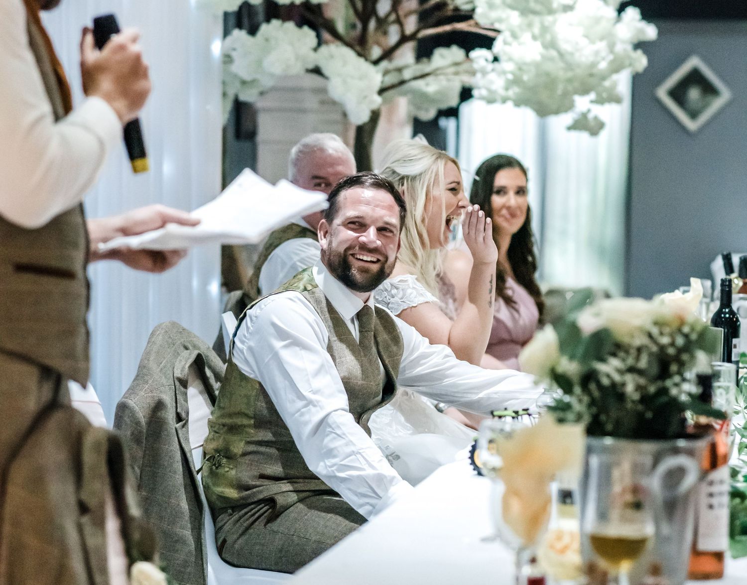 Wedding reception speech moment with decorated tables and white floral arrangements.