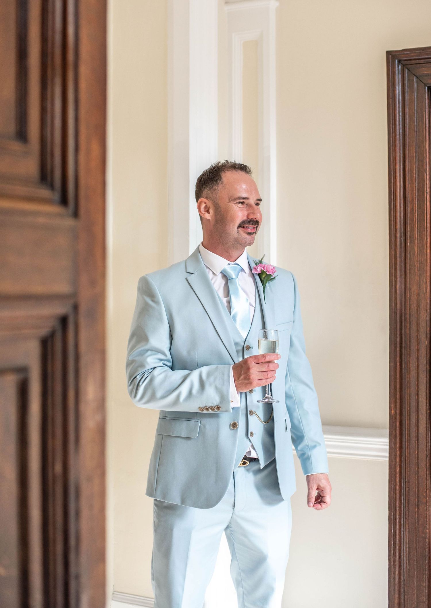 Wedding guest in pale blue suit holding champagne glass indoors.