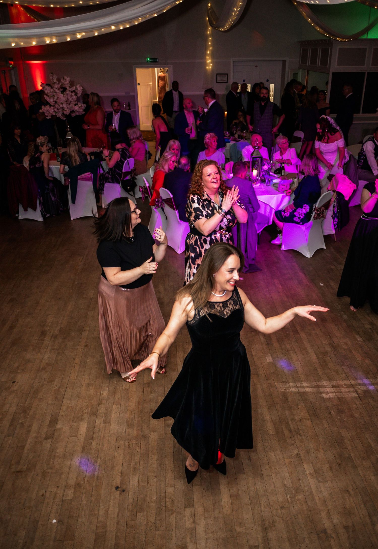 Wedding guests dancing under purple lighting at an evening reception with white chair covers.