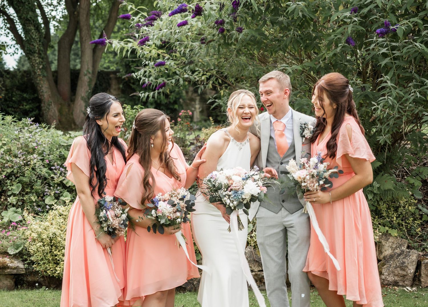 Wedding party in coral dresses and grey suit pose for photos in a garden setting.