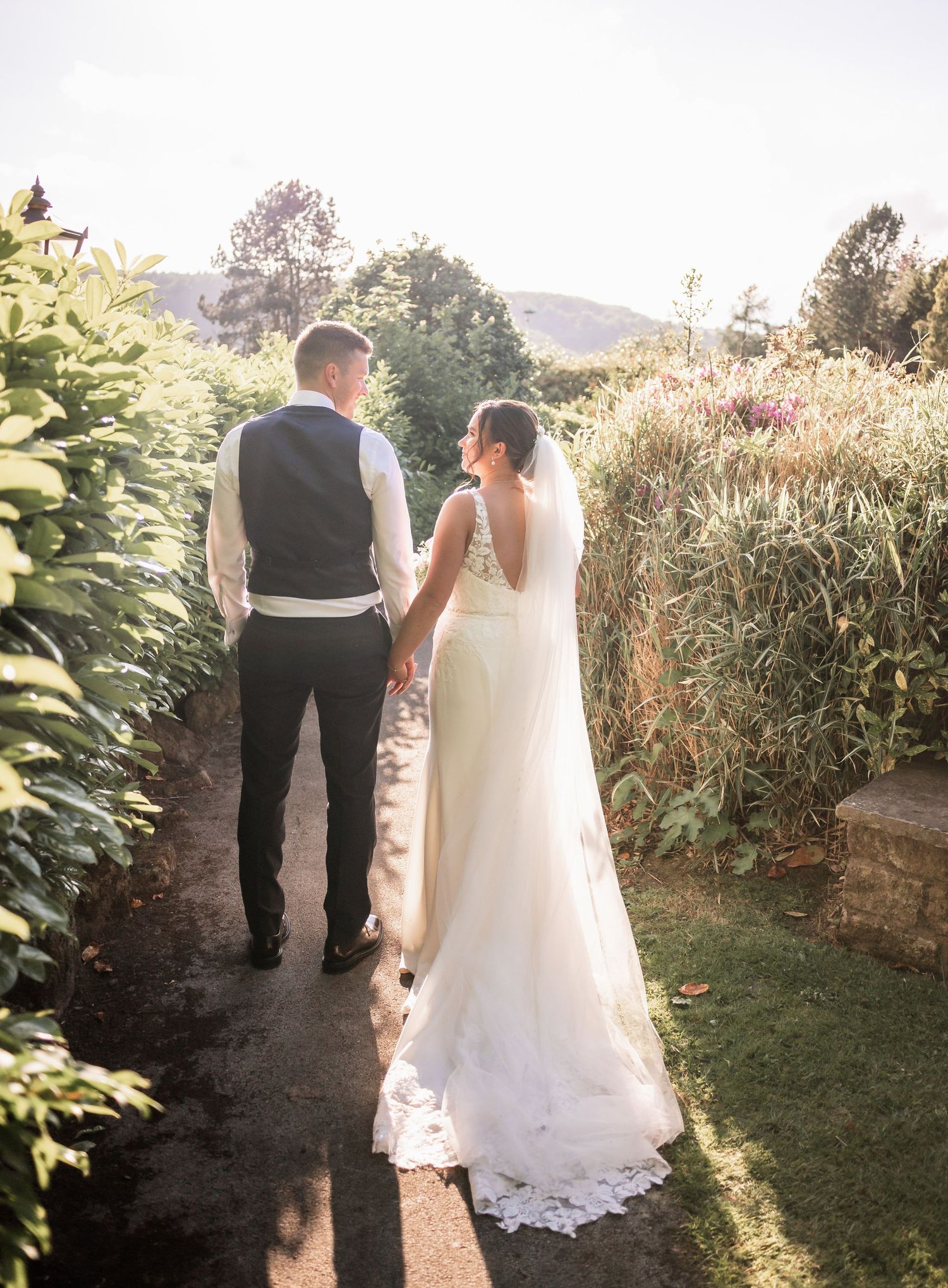 Newlyweds walk hand in hand down garden path at sunset overlooking scenic hillside.