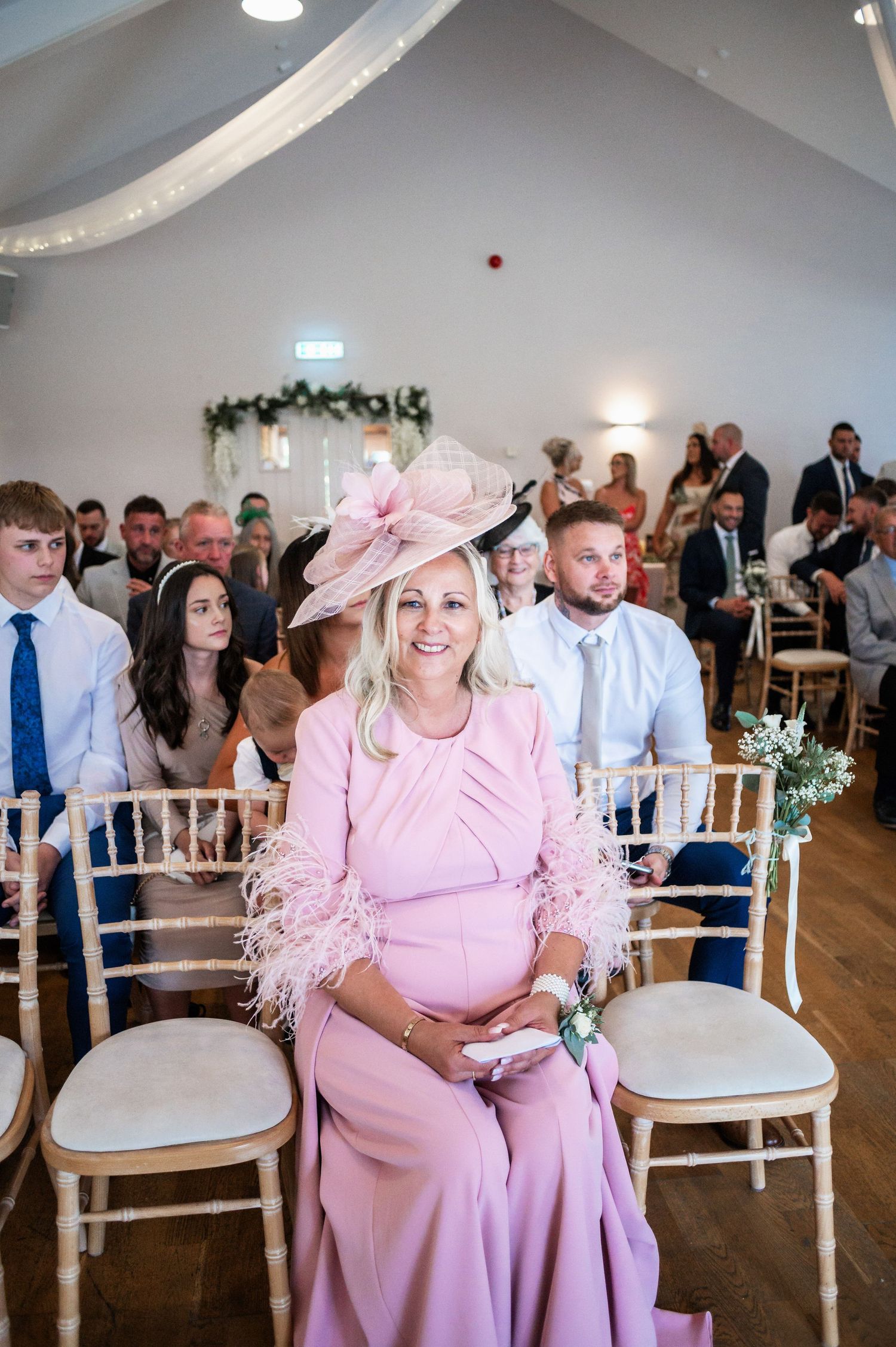Wedding guest in pink dress and matching hat seated in decorated ceremony venue.