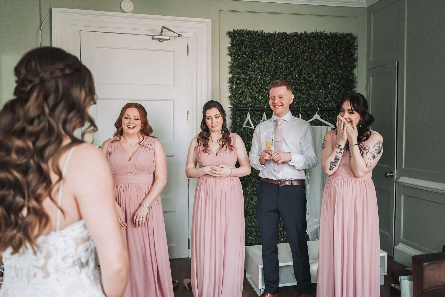 A bride reveals her dress to wedding party members in pink gowns in a green room.