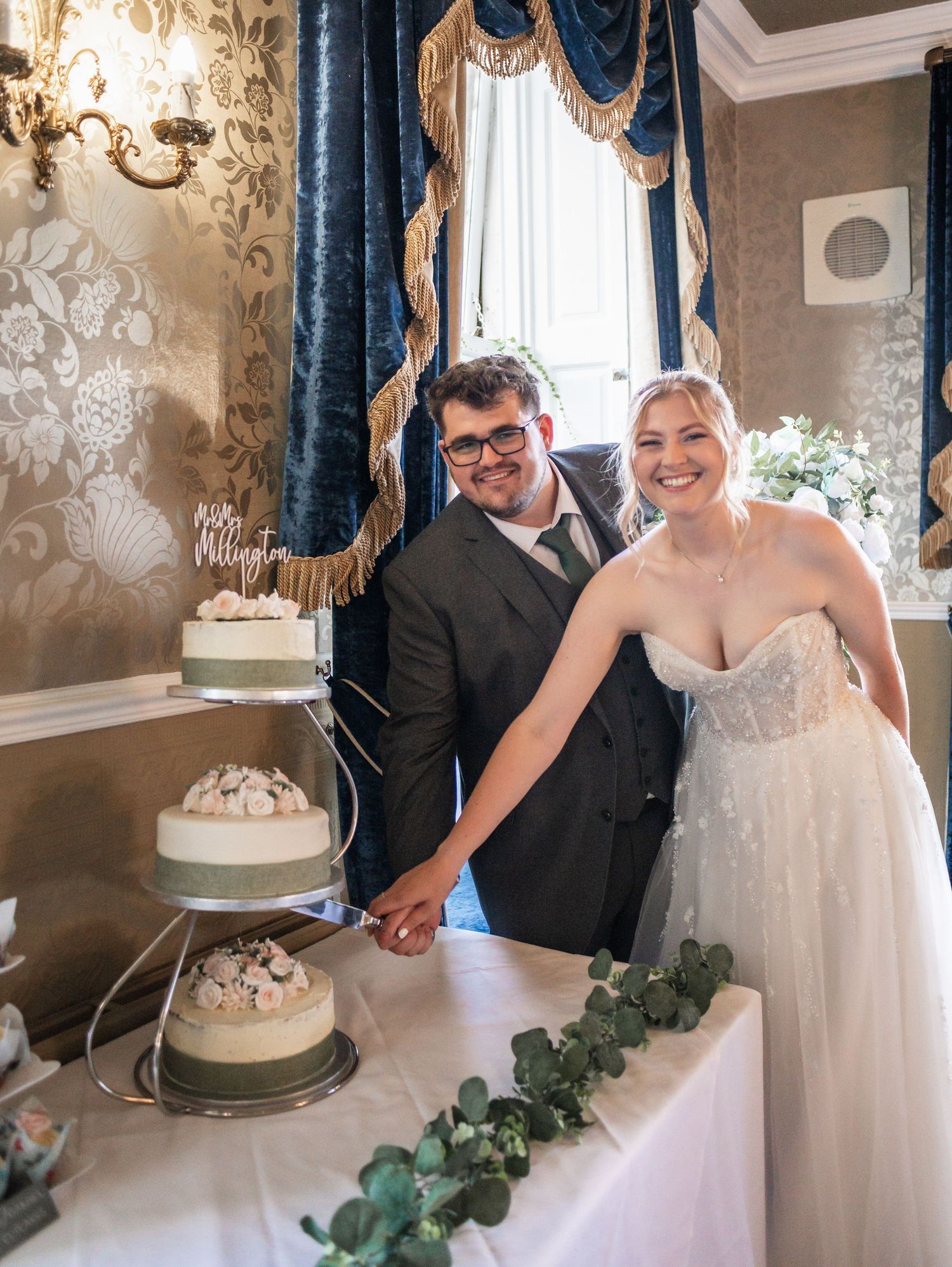 Wedding couple cuts their white tiered cake in elegant room with blue drapes and ornate wallpaper.