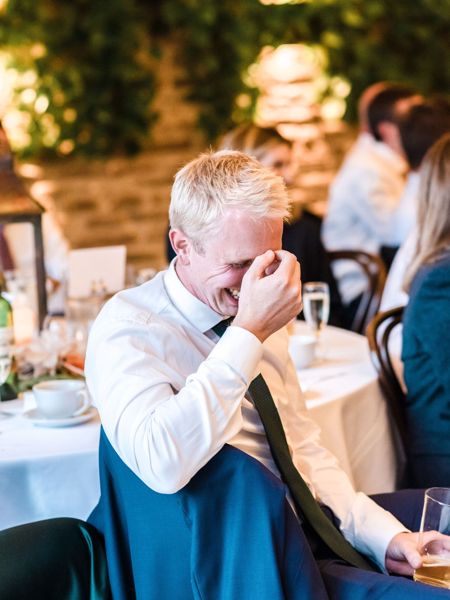 Wedding guest laughs with hand covering face during reception speech.