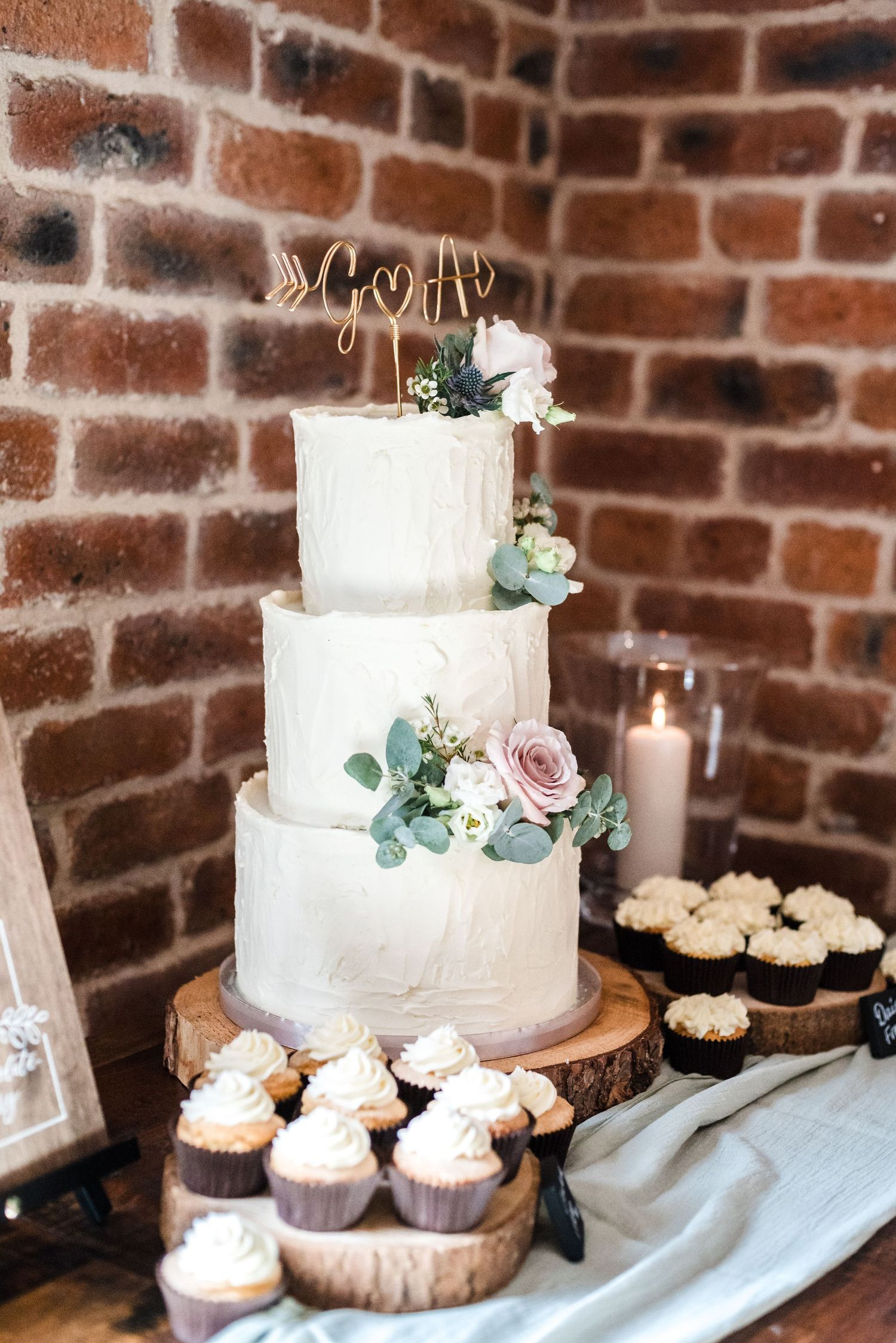 Three-tiered white wedding cake with roses and eucalyptus on wooden stand with cupcakes.
