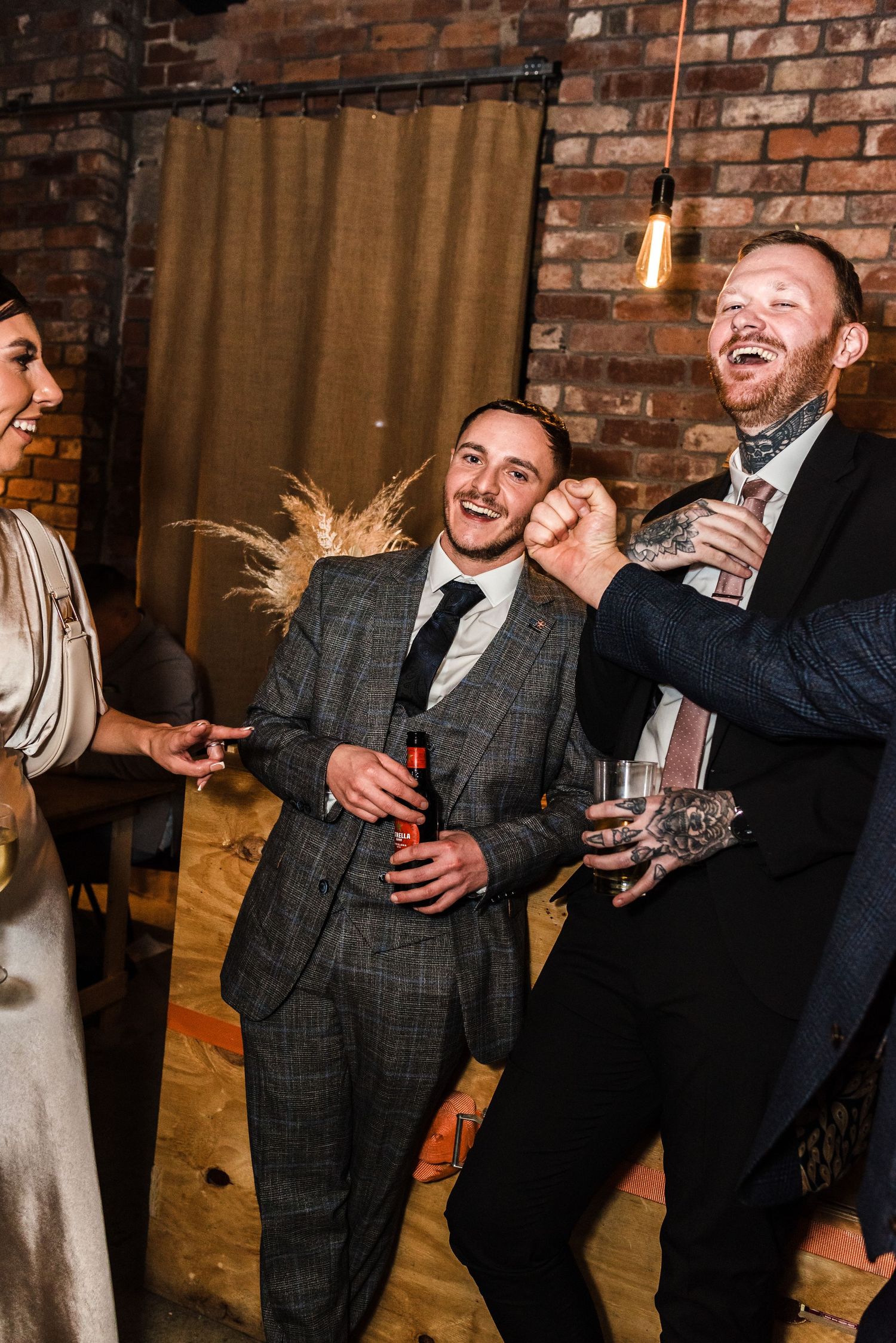 Wedding guests sharing laughs during reception in industrial-style venue with brick walls.