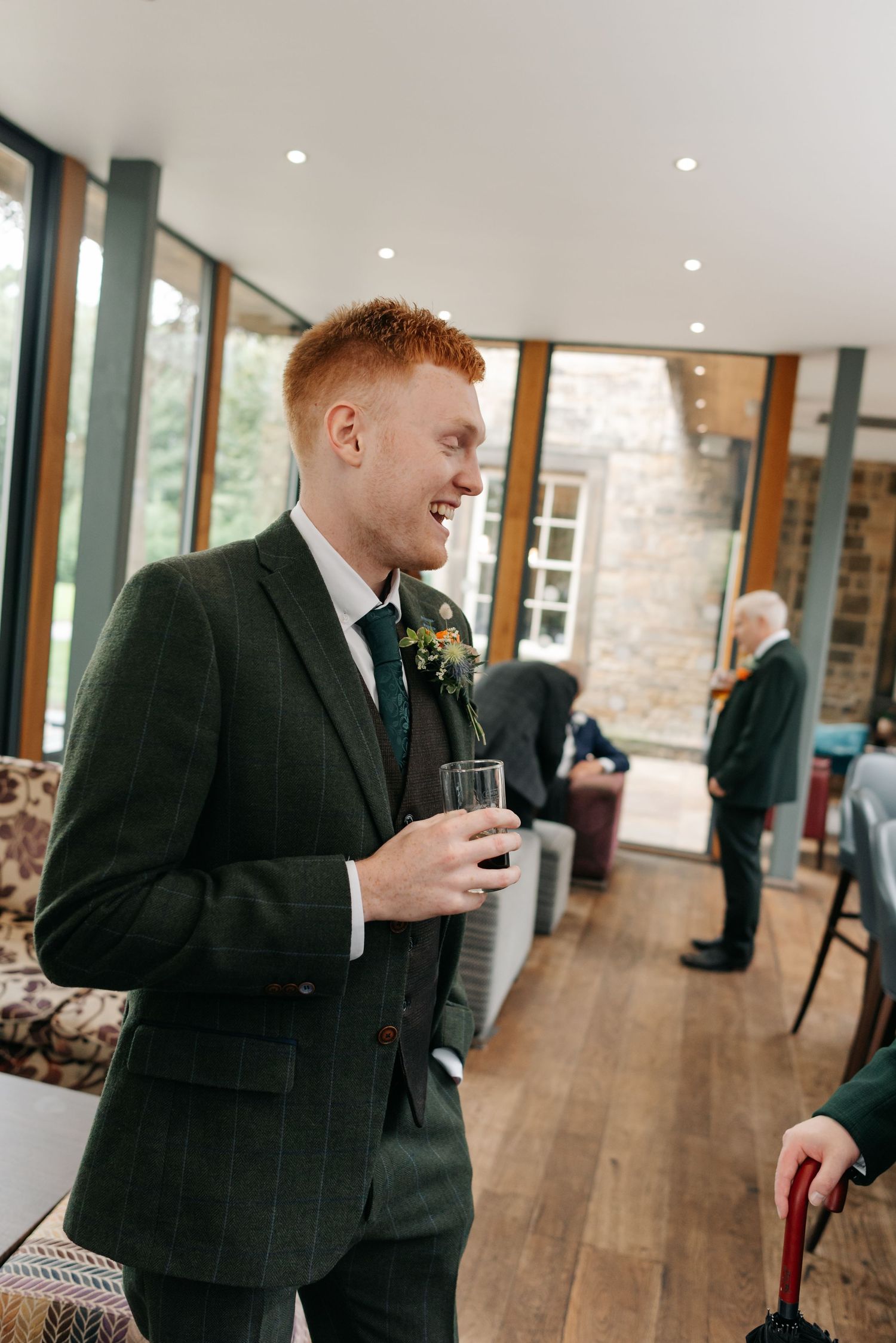 Wedding guest in a dark suit enjoying a drink in a modern venue with wooden floors.