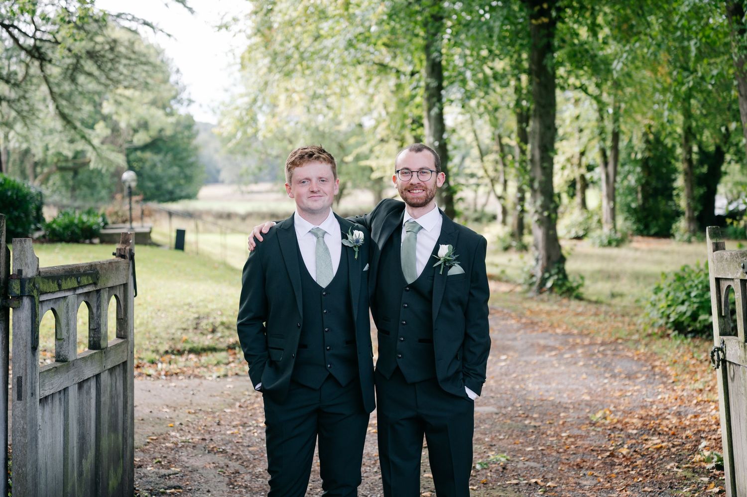 Two people in formal black suits stand together smiling outdoors in a wooded park setting.
