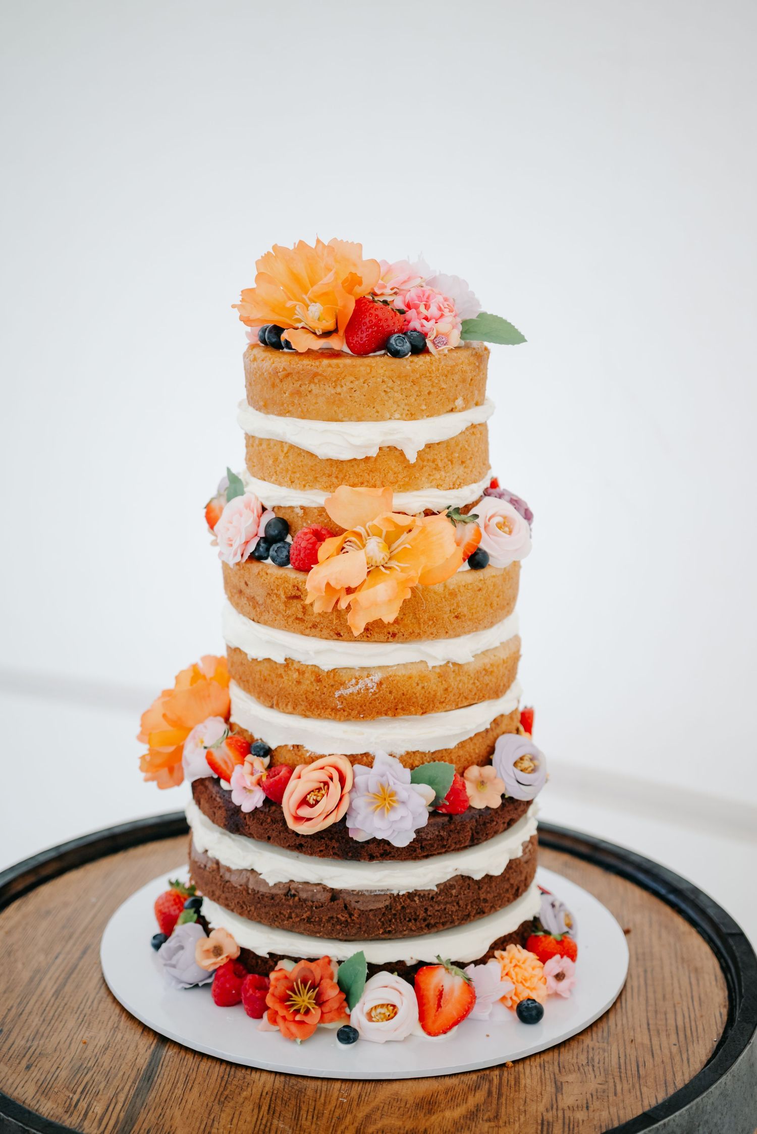 A tiered naked wedding cake decorated with fresh flowers and berries in orange, pink and red tones.