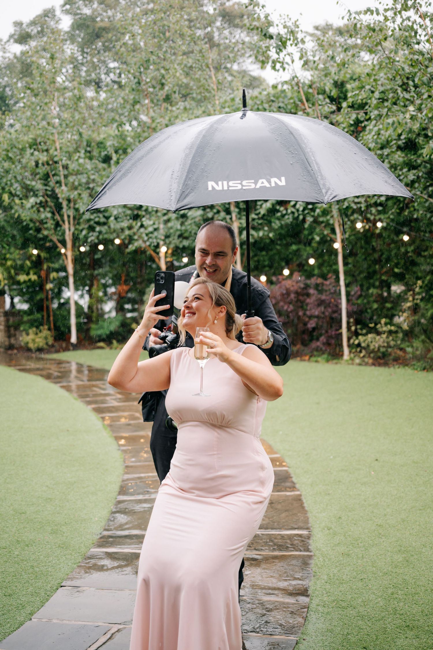 Wedding guests taking selfies with umbrellas during an outdoor celebration.