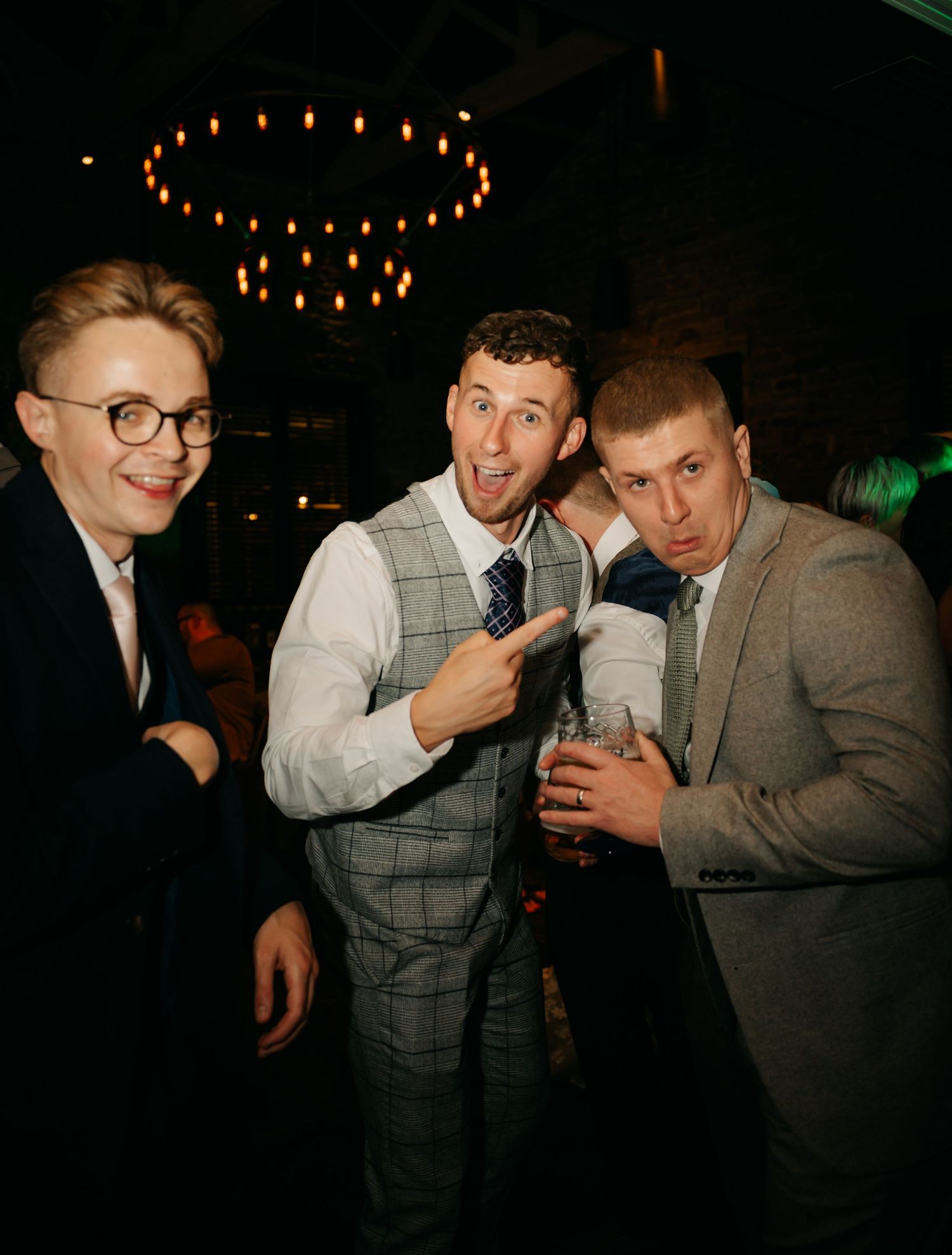 Three wedding guests pose together under moody lighting with a circular chandelier overhead.