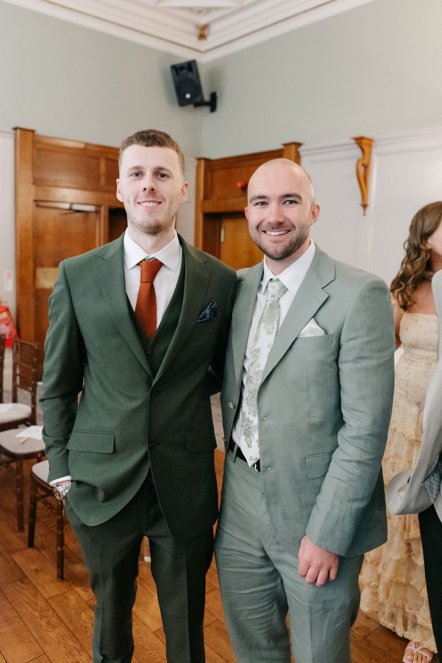Groomsmen wearing matching sage green suits with burgundy ties stand together in a bright indoor venue.