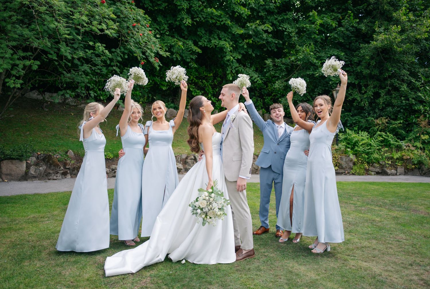 Wedding party in light blue dresses and white gowns pose on green lawn.