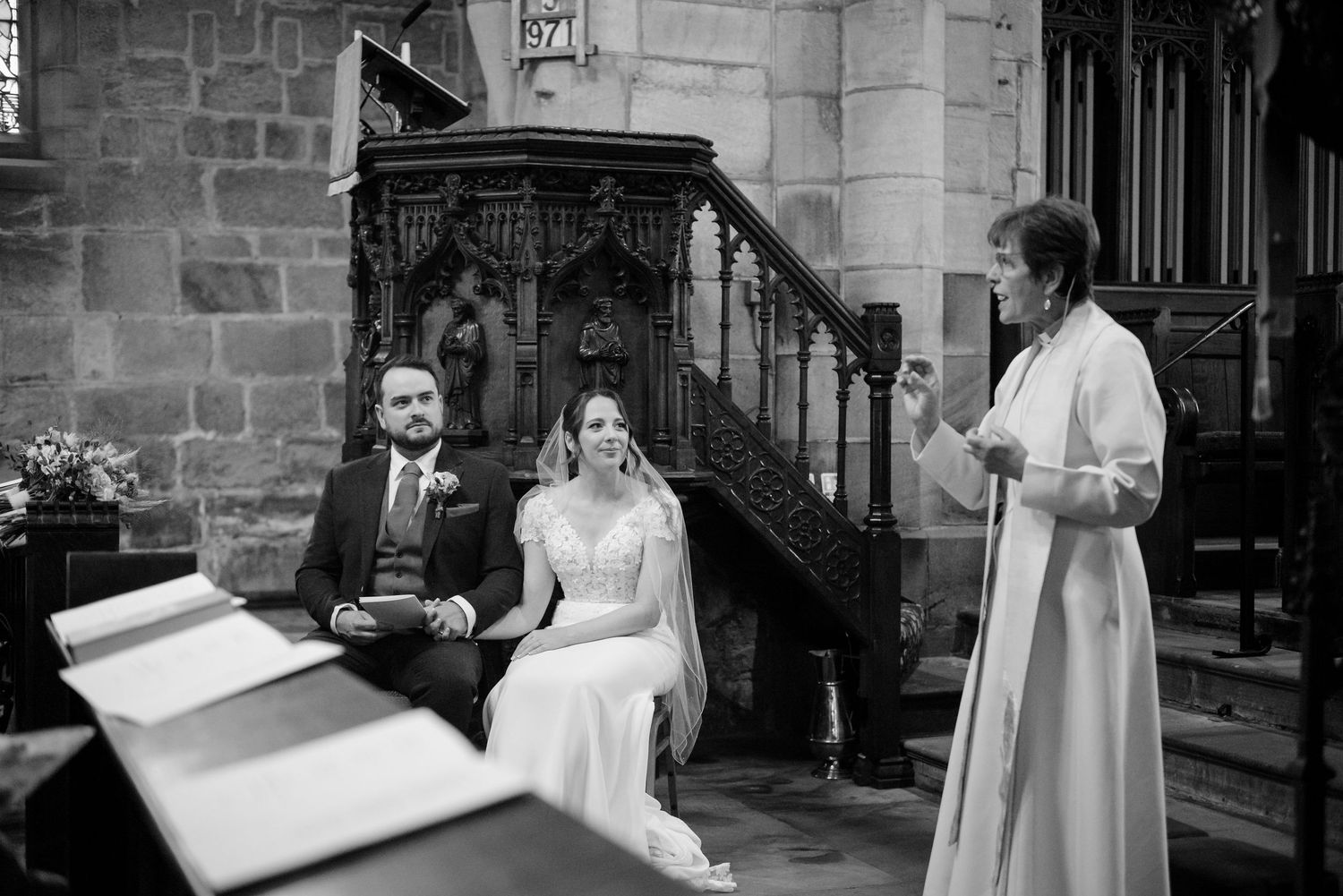 Bride and groom sit side by side in a stone church as an officiant in white robes leads the wedding ceremony.