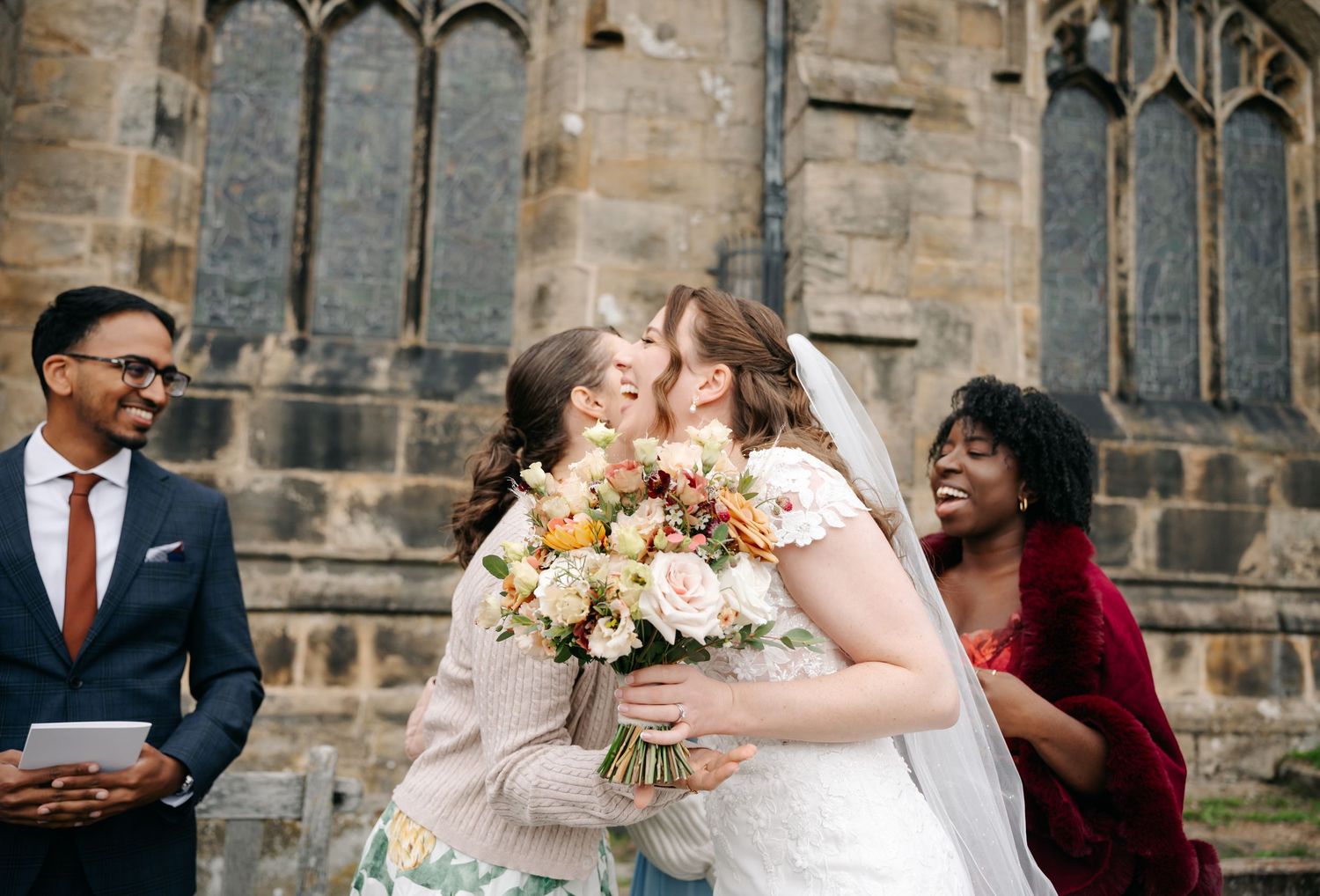 A bride in a white lace gown and veil embraces a guest holding a bouquet outside a stone church as other guests smile nearby.