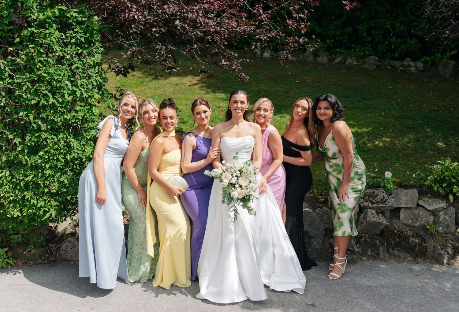 Bridesmaids in pastel and purple dresses pose together outdoors in a garden setting.