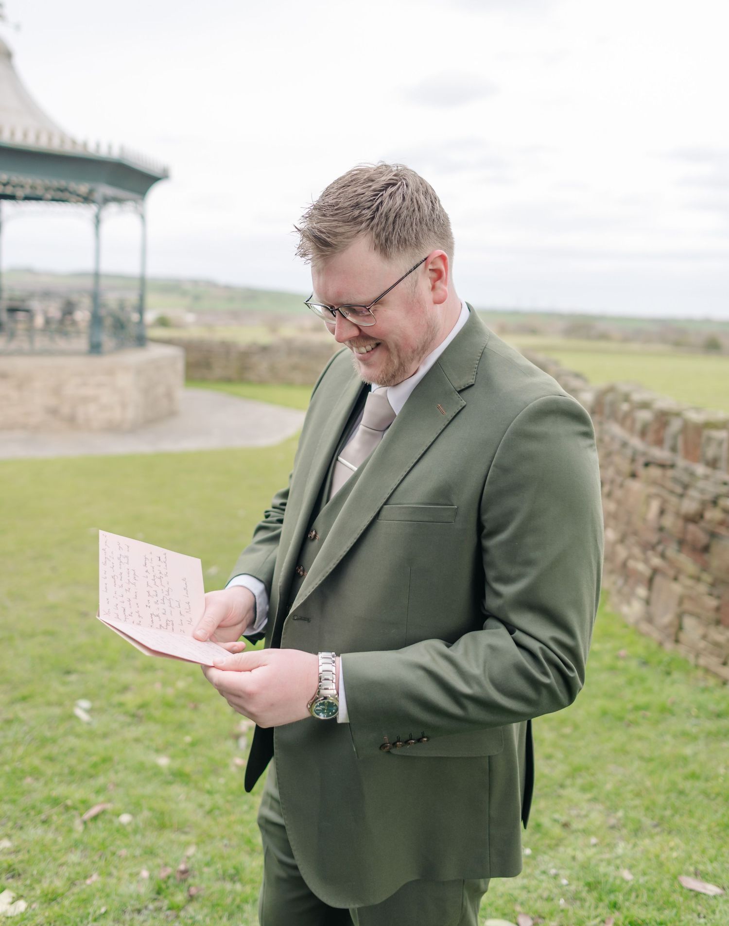 A person in a green suit jacket reads a paper while standing outdoors near a gazebo.