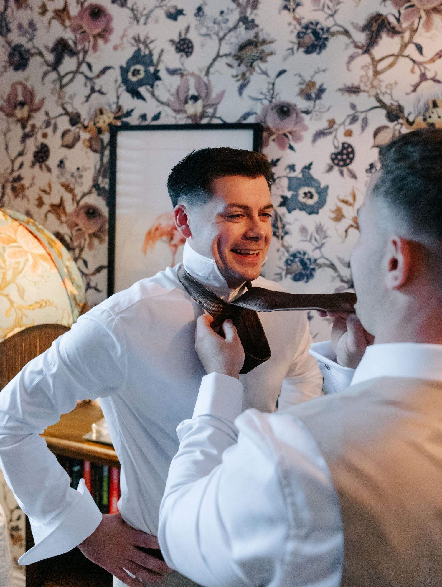 Two people in white shirts share a playful moment while adjusting clothing in a room with floral wallpaper.