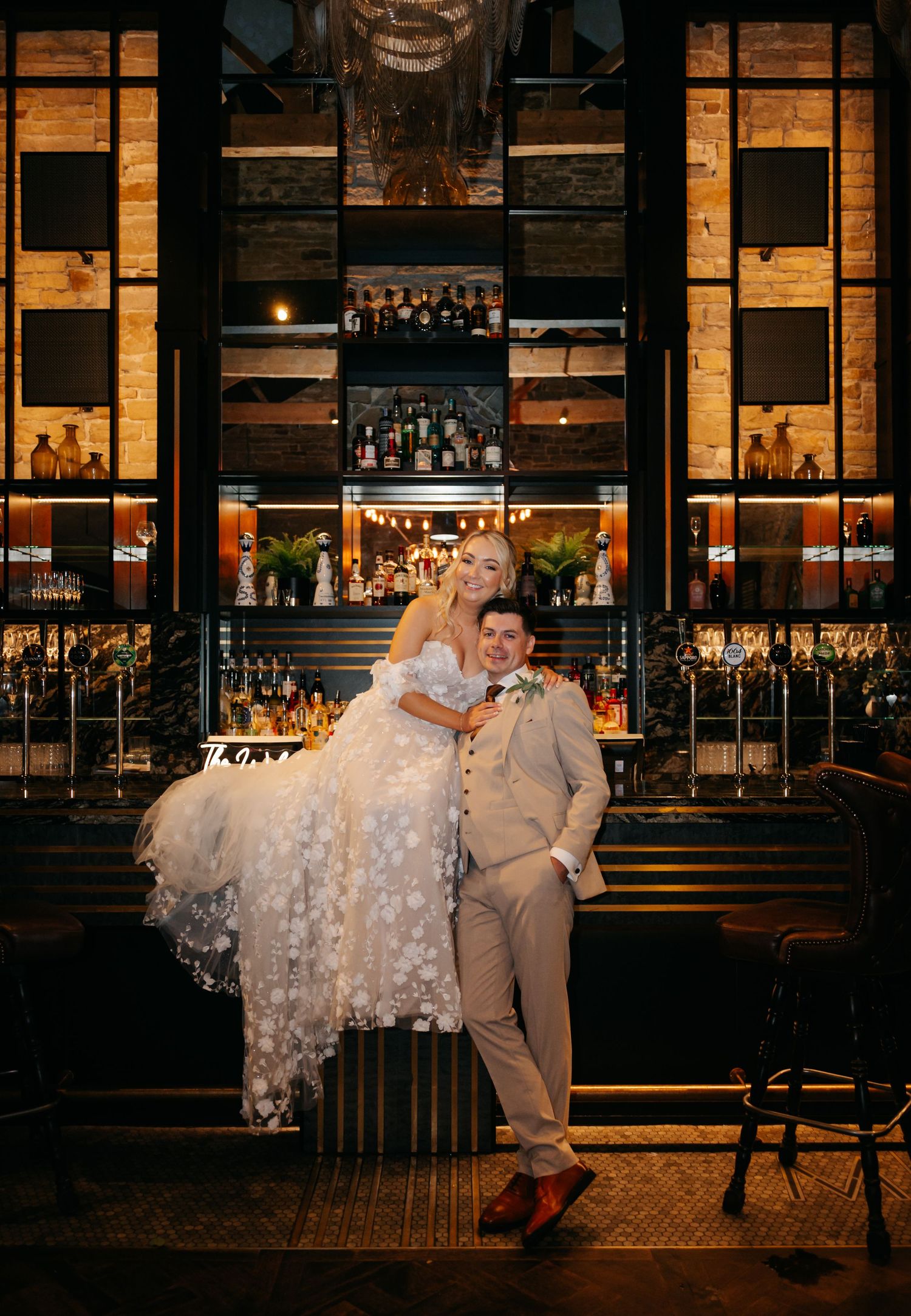 A wedding couple poses against an elegant bar backdrop with illuminated shelves of bottles and warm lighting.
