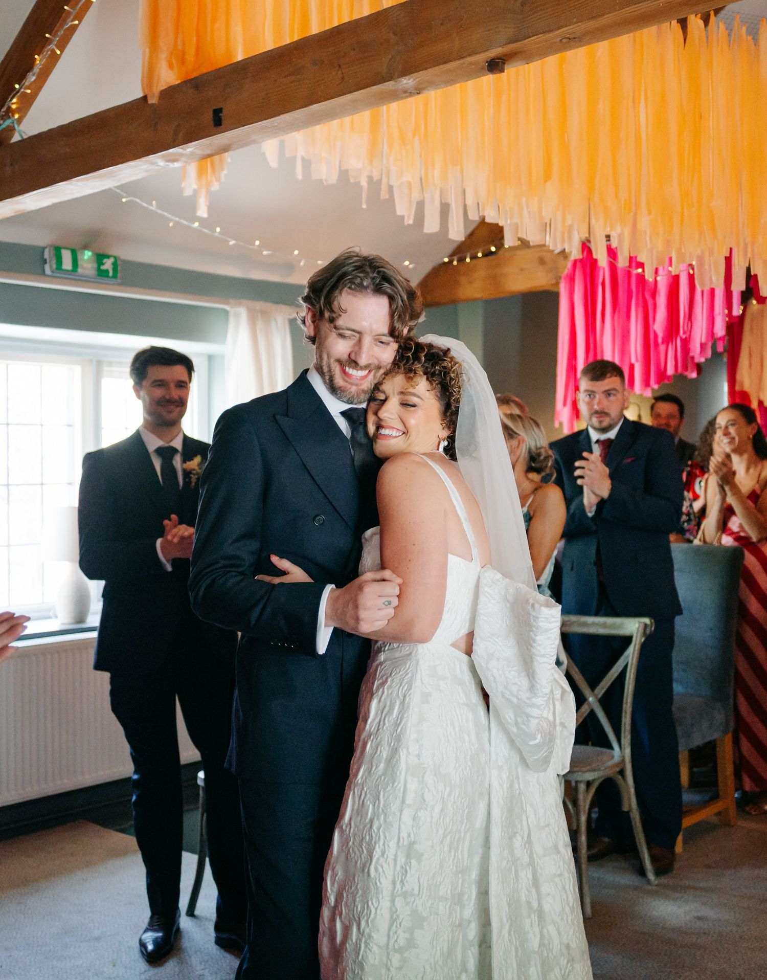 Wedding couple celebrates with guests under festive pink and orange streamers.