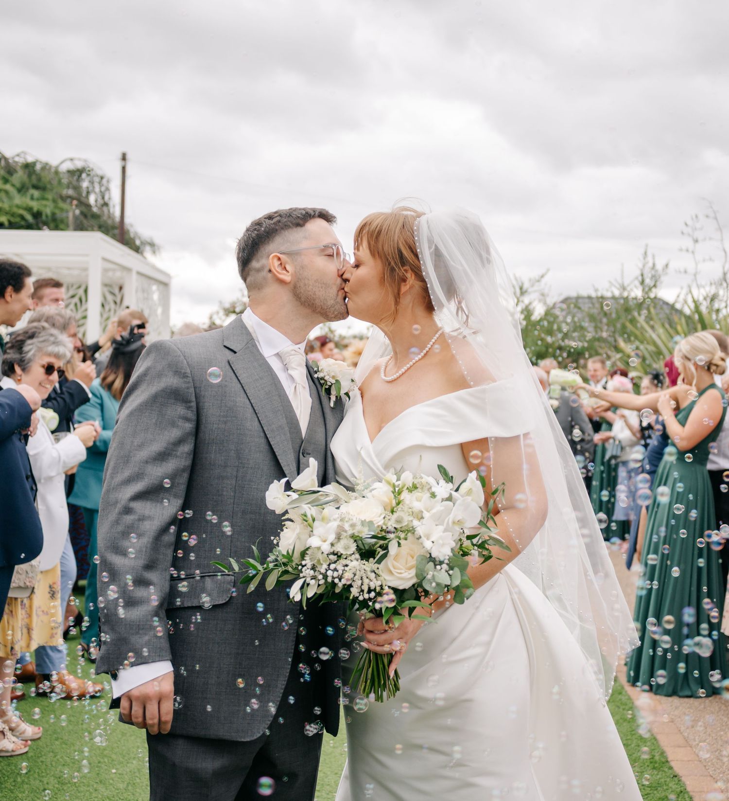 Wedding couple shares a kiss outdoors while guests celebrate with confetti.