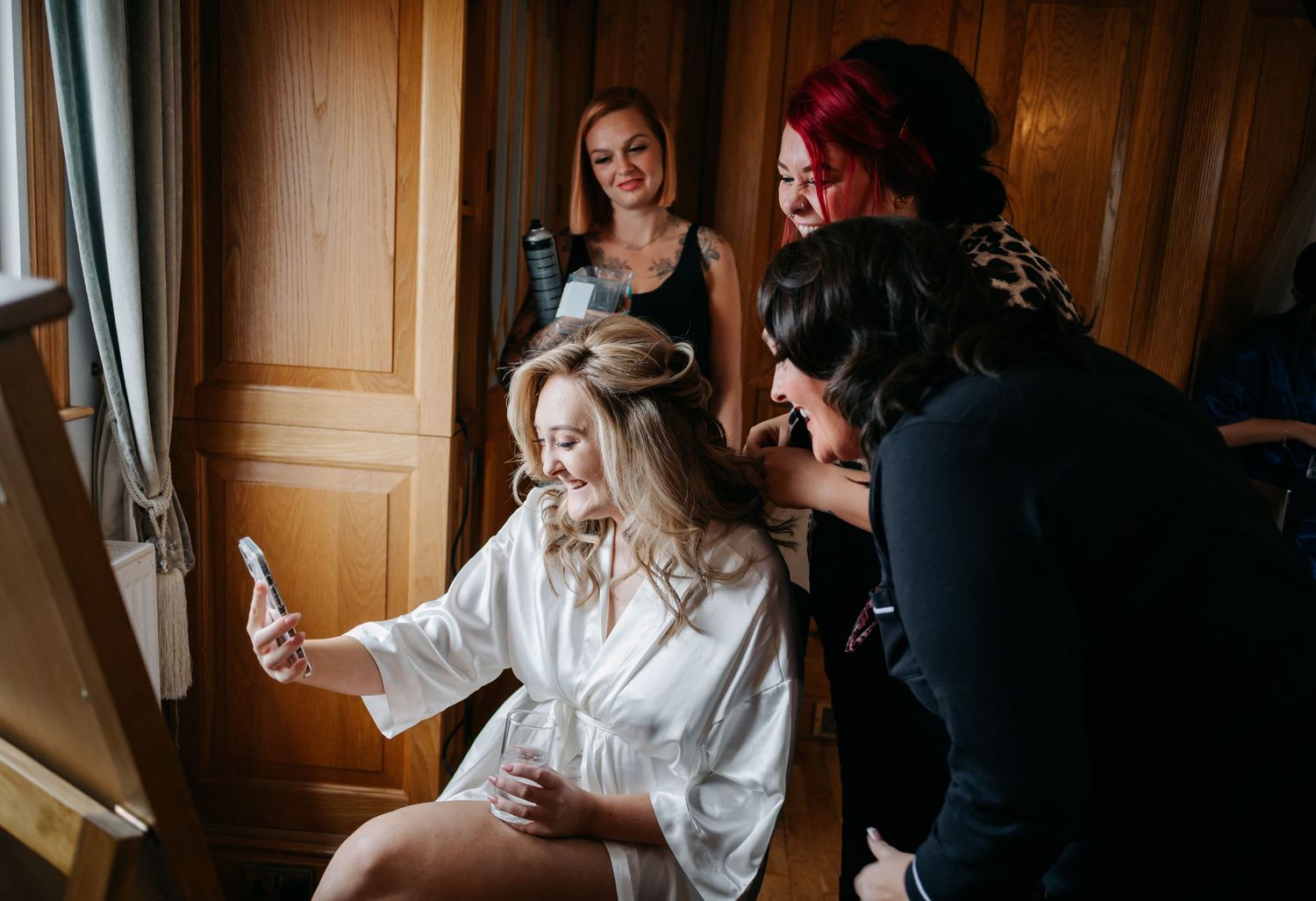 Wedding party laughs together during hair and makeup preparations in a warmly lit hotel room.