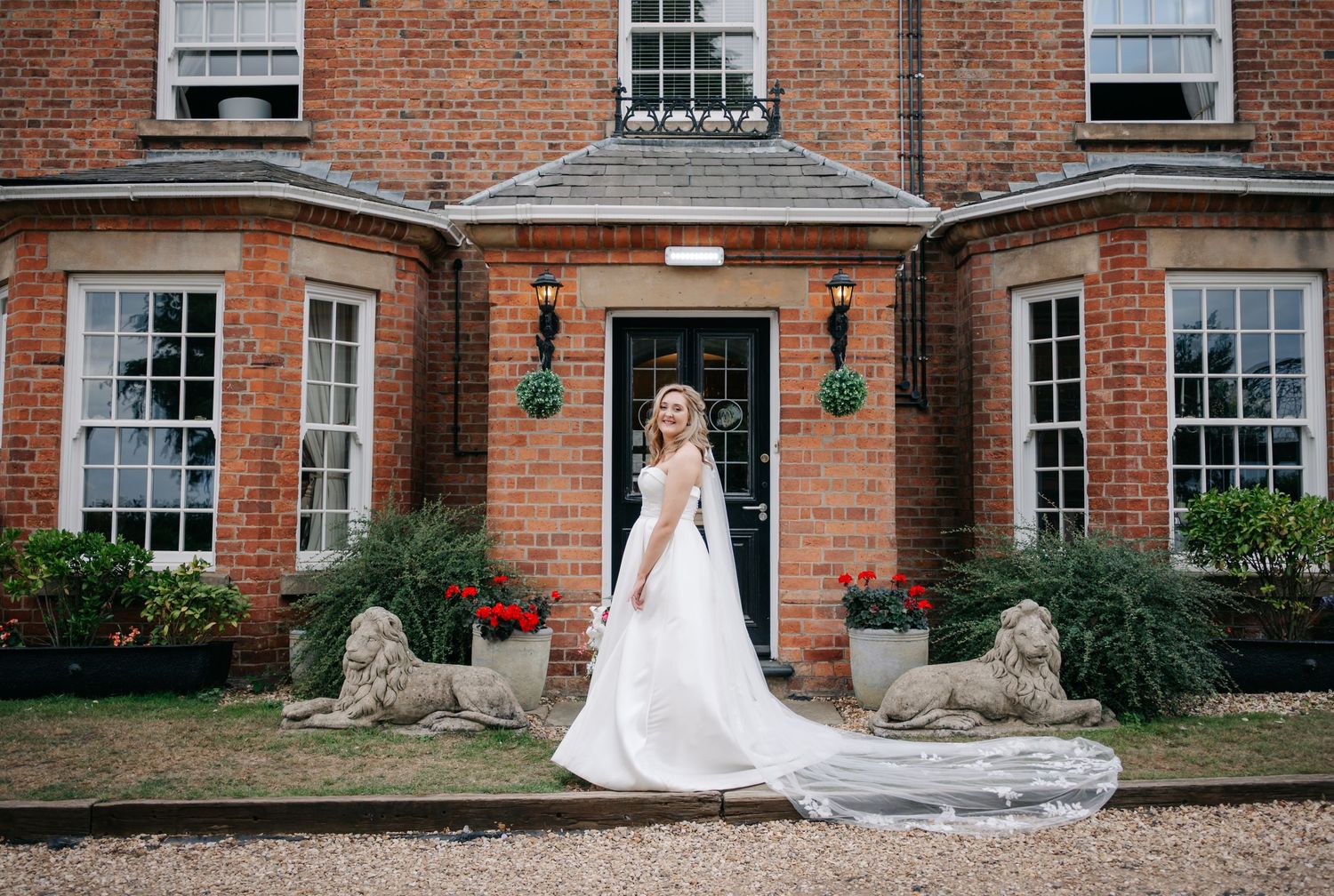Elegant bride in white gown and veil poses in front of red brick mansion with stone lion statues.