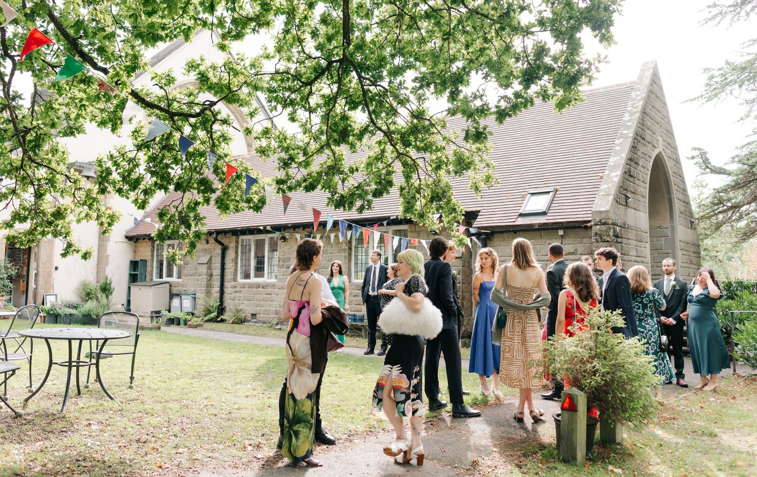 Wedding guests gather outside a charming stone chapel during a garden reception with trees and flowers in bloom.