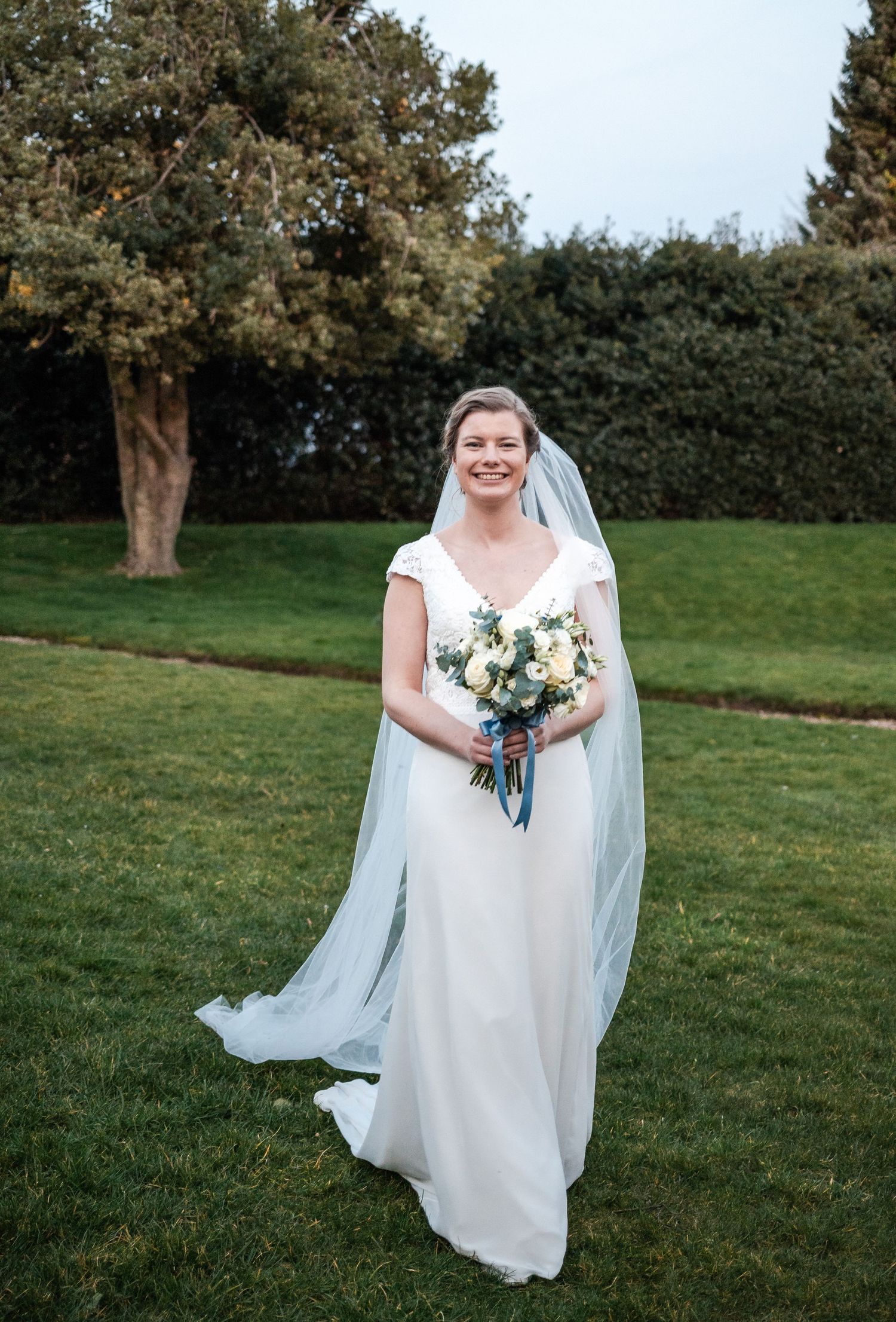 A bride in a flowing white wedding dress holds a blue and white bouquet while standing on a grassy lawn.