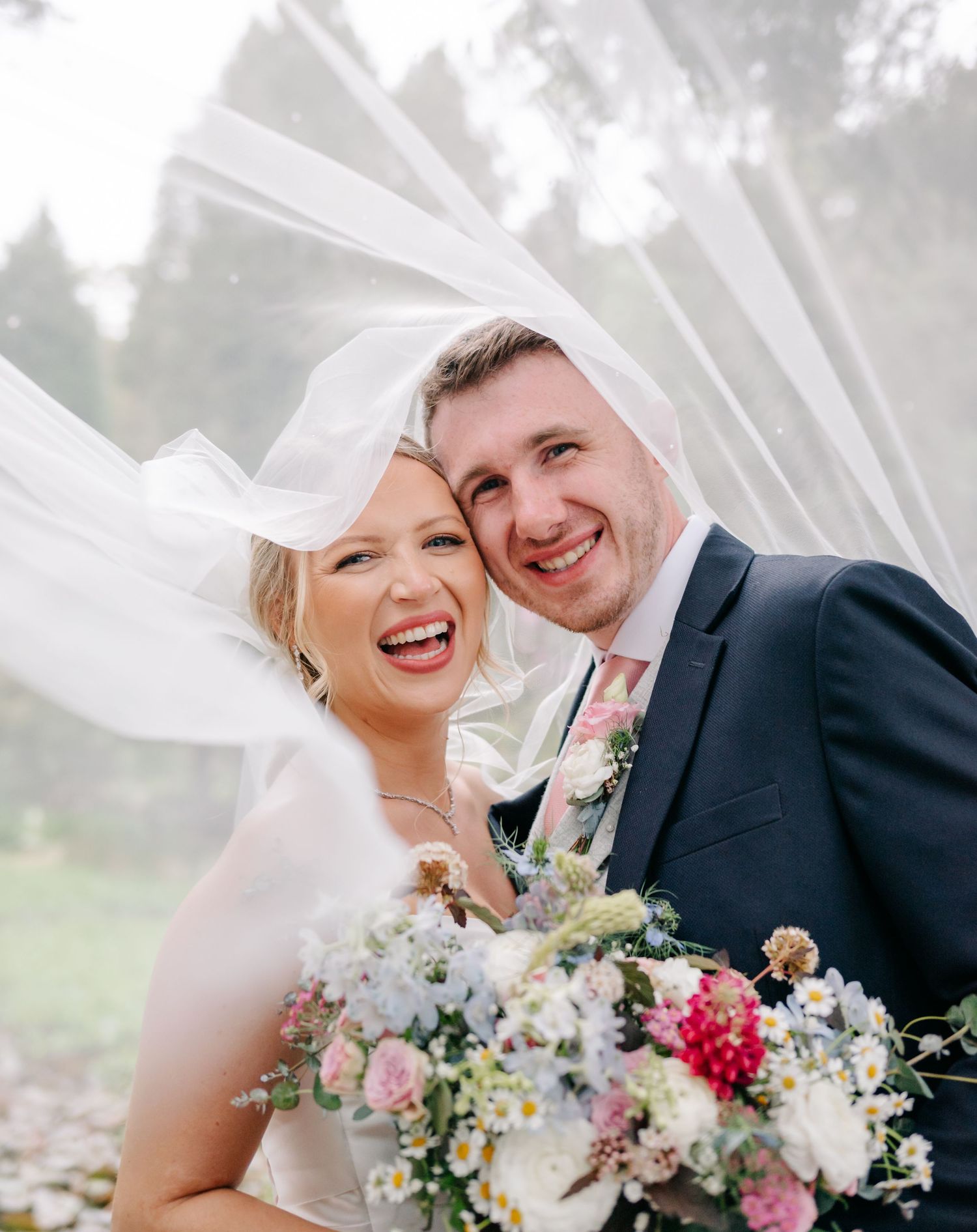 A joyful wedding couple shares a candid moment while holding a colorful spring bouquet of flowers outdoors.