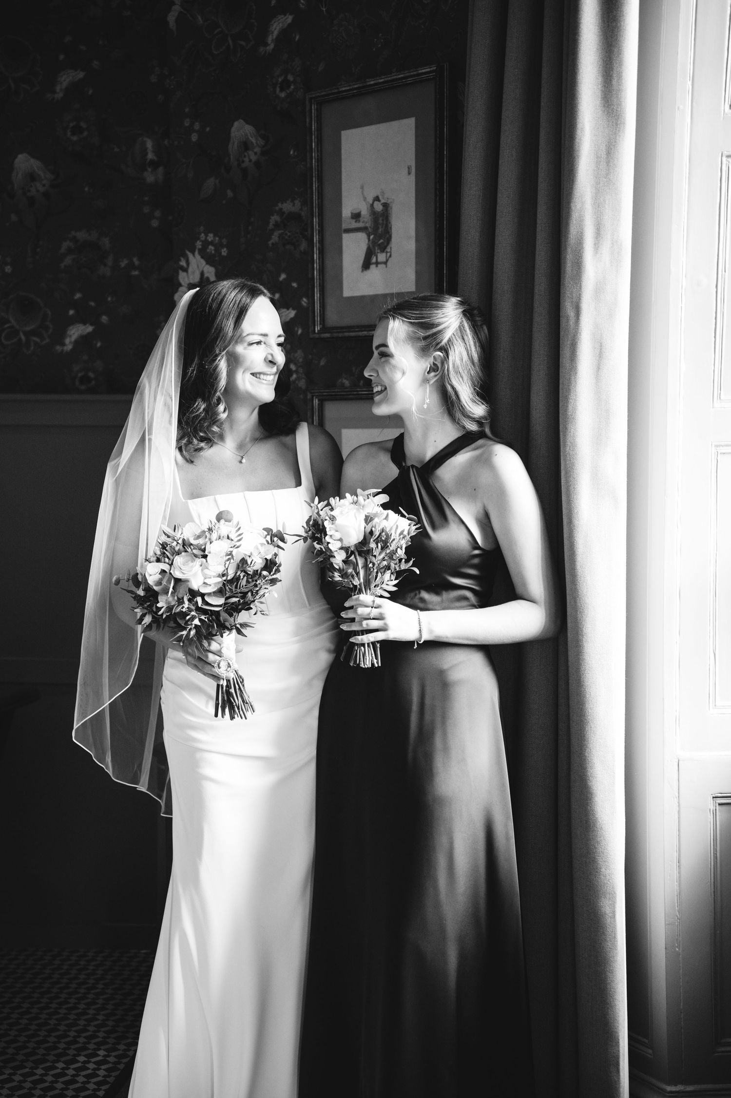 Black and white photo of a smiling bride in a veil and bridesmaid in a halter gown holding bouquets indoors.
