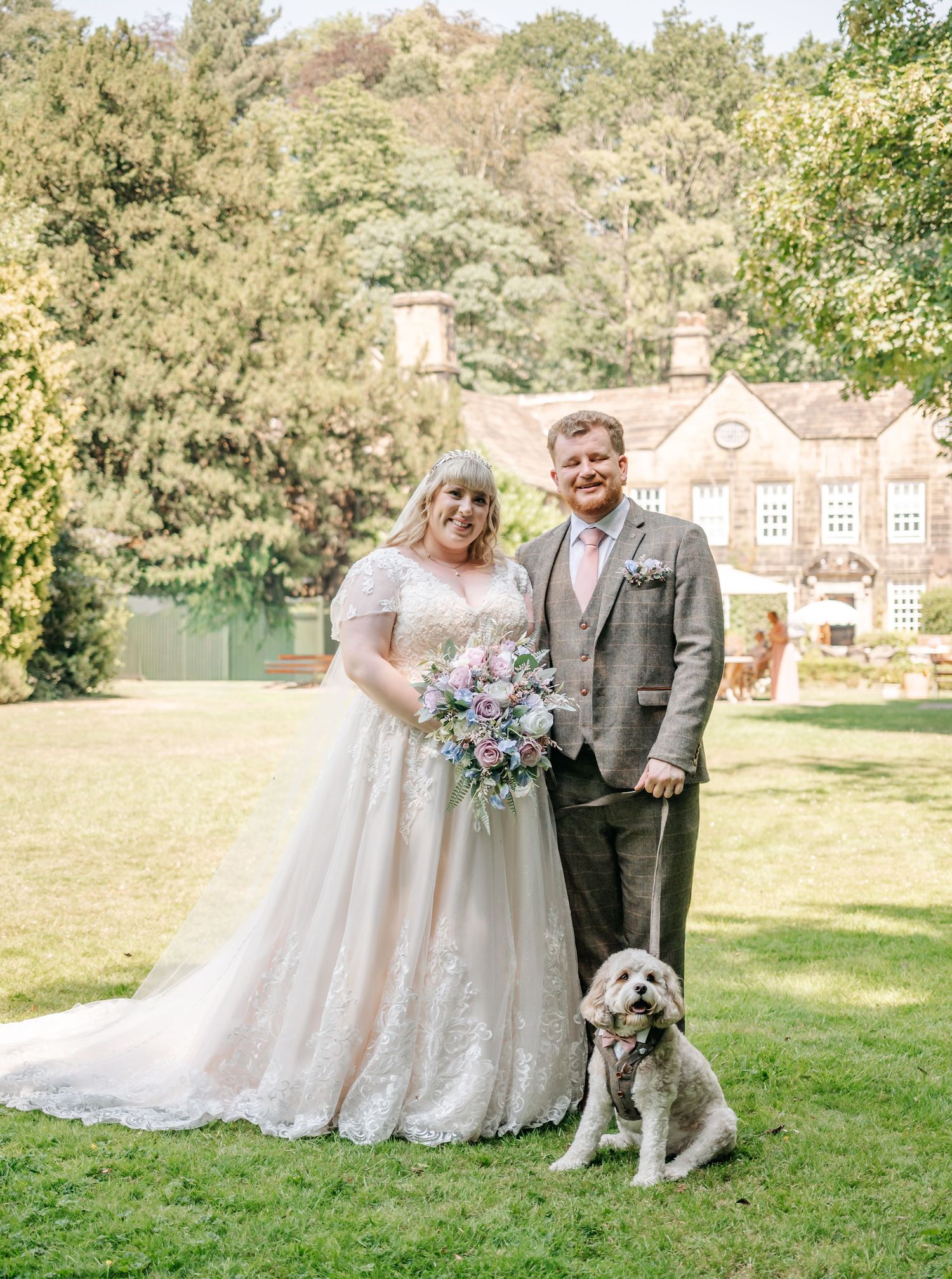 Smiling bride and groom pose with their dog in front of a charming English manor on their wedding day.