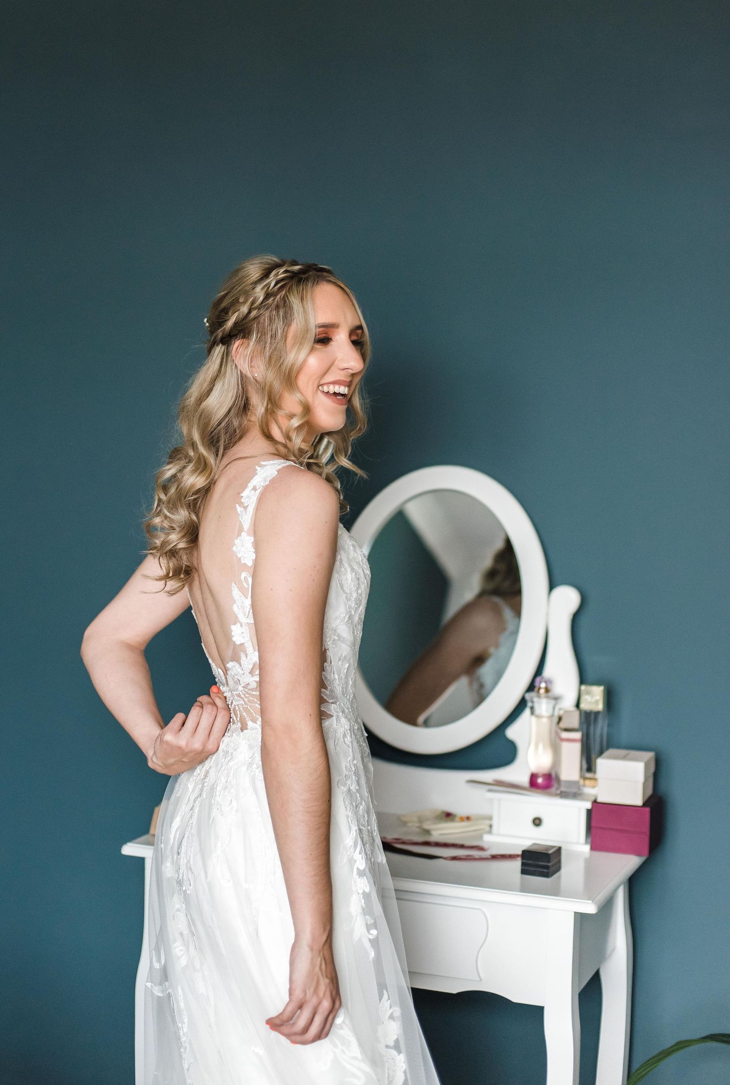 Elegant bride in lace wedding dress poses next to white vanity mirror in teal room.