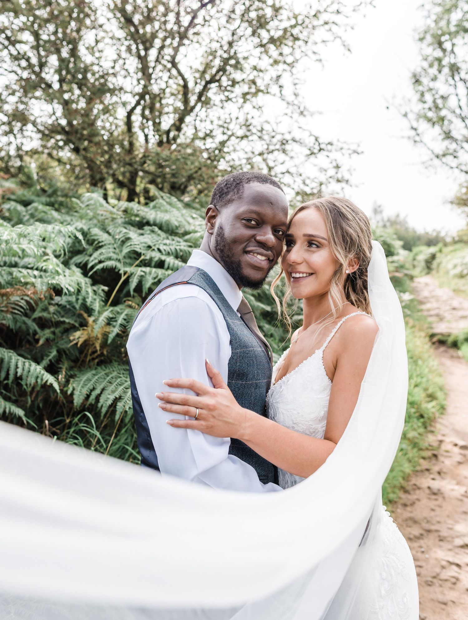 A newlywed couple embraces outdoors surrounded by lush greenery and ferns.