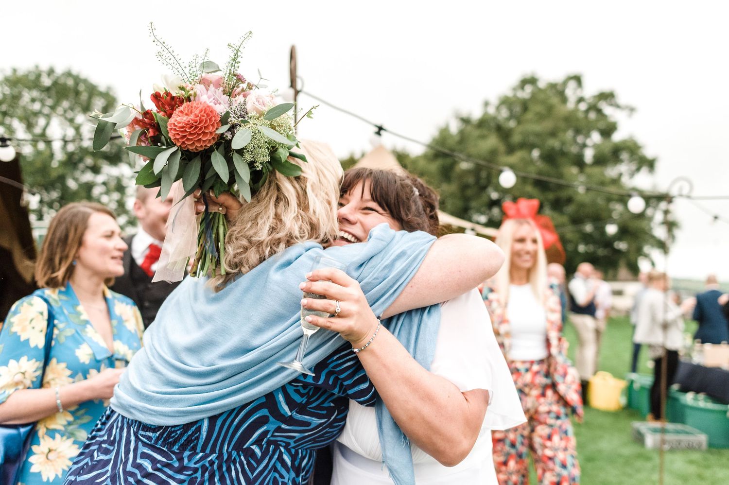 Joyful celebration at an outdoor wedding with guests hugging and holding a floral bouquet.