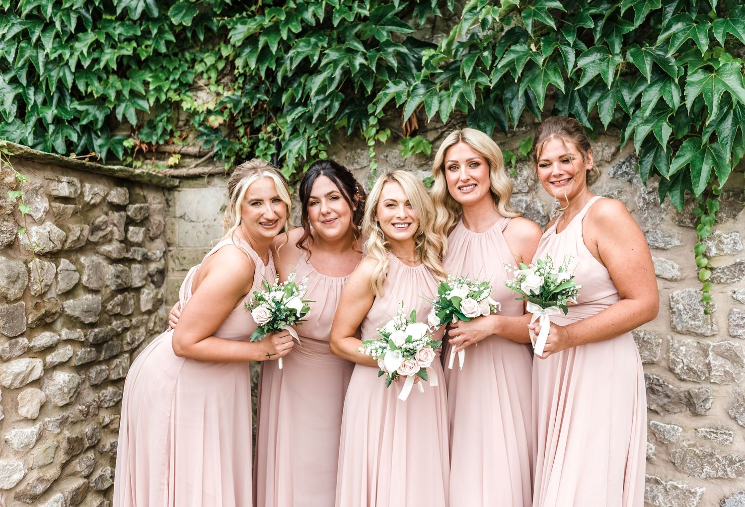 Wedding party in blush pink dresses pose together against a stone wall with floral bouquets.