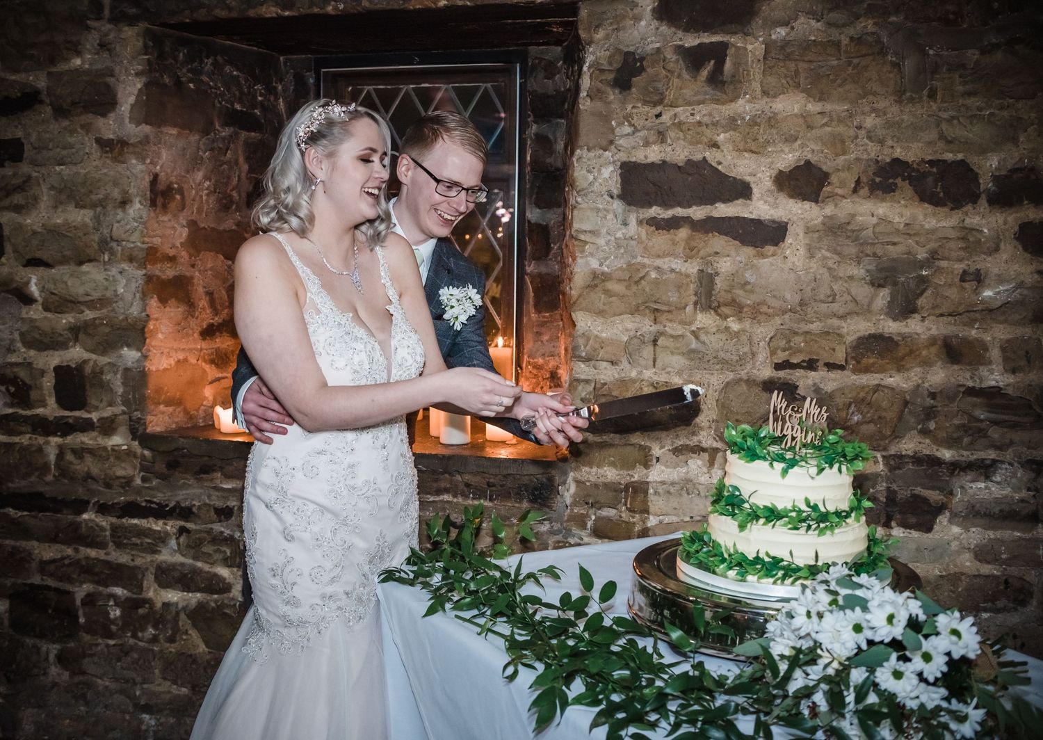 A couple cuts their green and white wedding cake together against a rustic stone wall backdrop.
