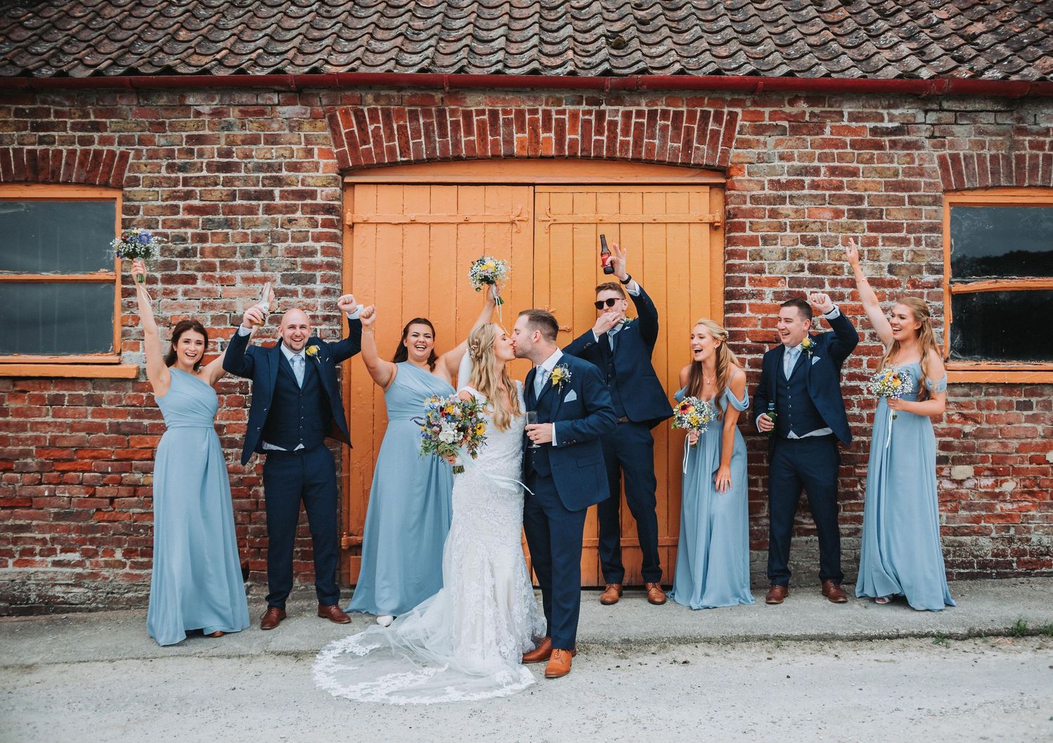 Wedding party in blue bridesmaid dresses poses against rustic brick barn.