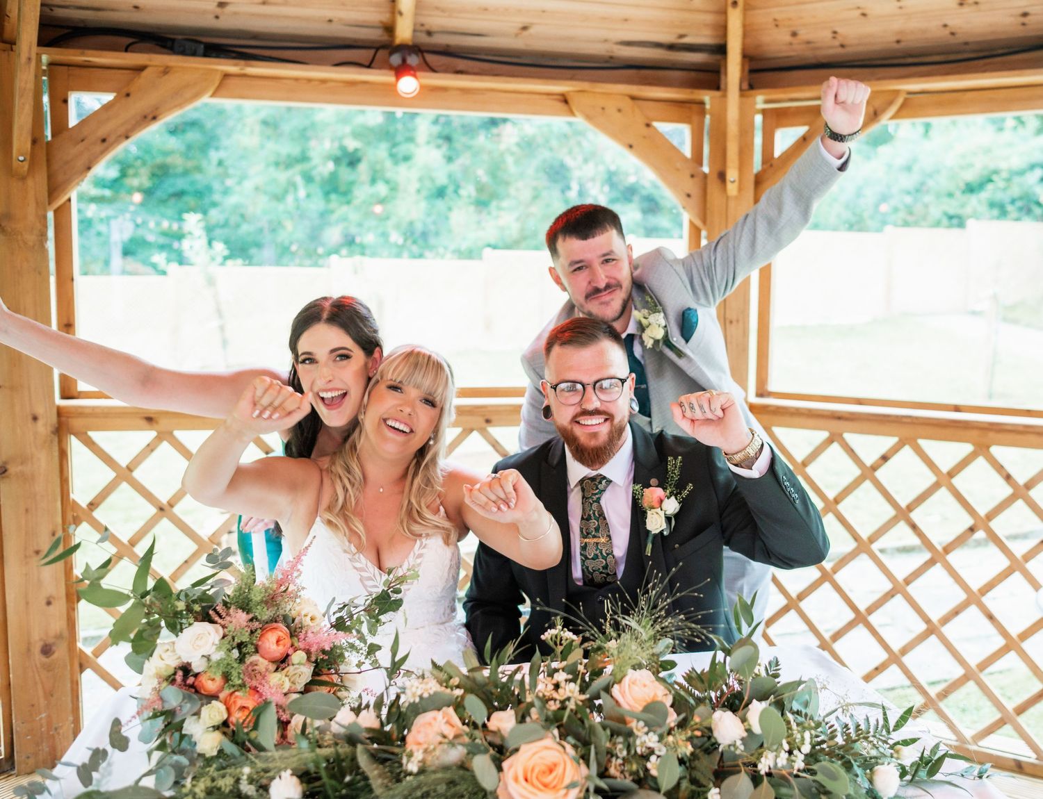Wedding party poses joyfully together at wooden gazebo with floral decorations and string lights.