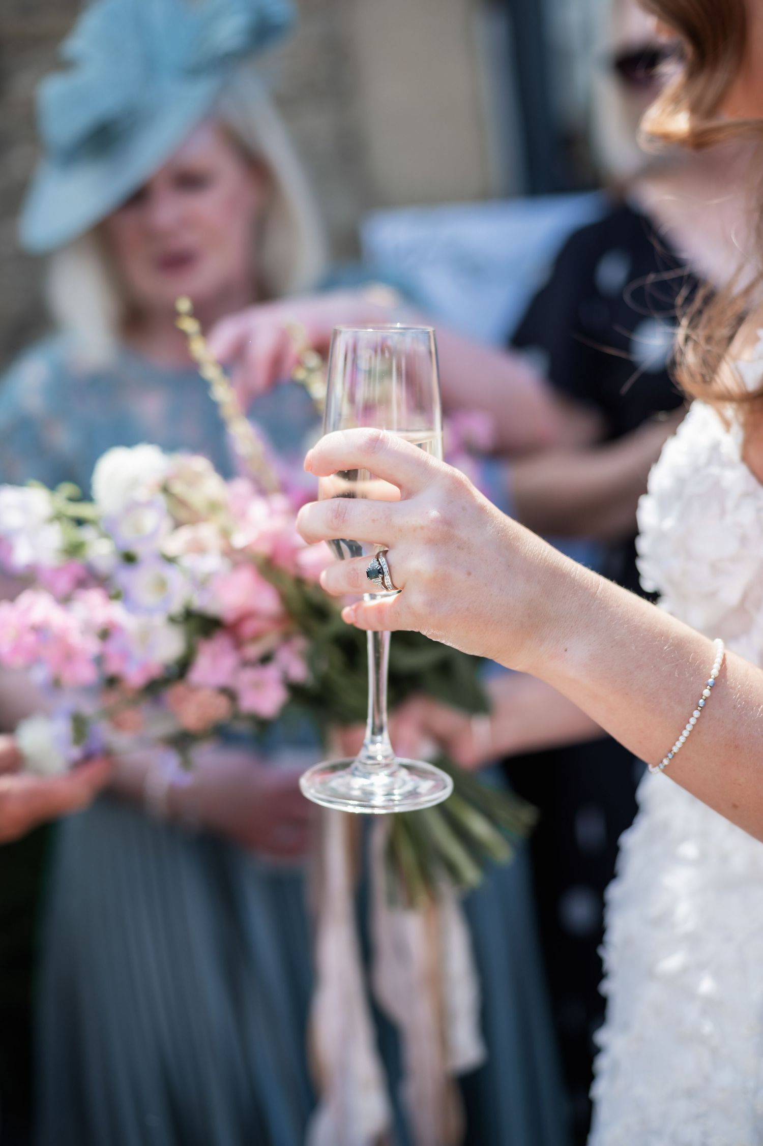 An elegant champagne glass is held by a hand wearing delicate jewelry with pink floral arrangements in the background.
