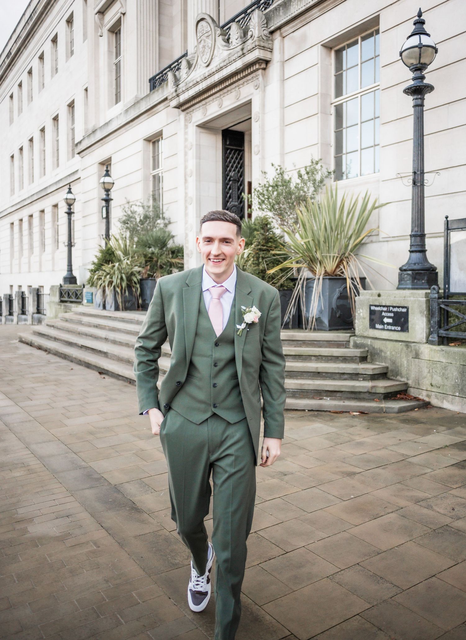 A wedding guest wearing a sage green three-piece suit and sneakers walks outside a classic stone building.