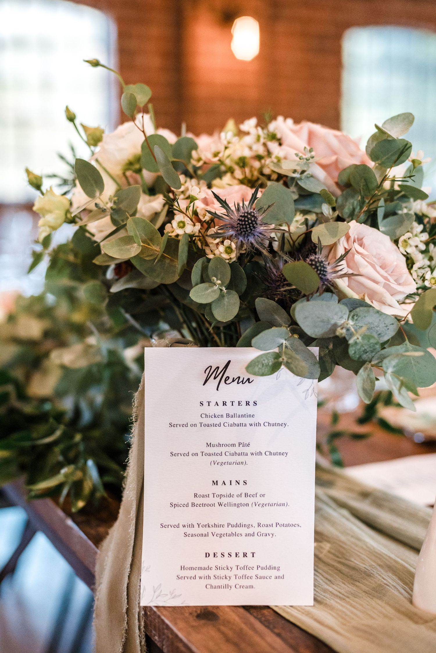 Elegant wedding menu card displayed with blush pink roses and eucalyptus on wooden table.