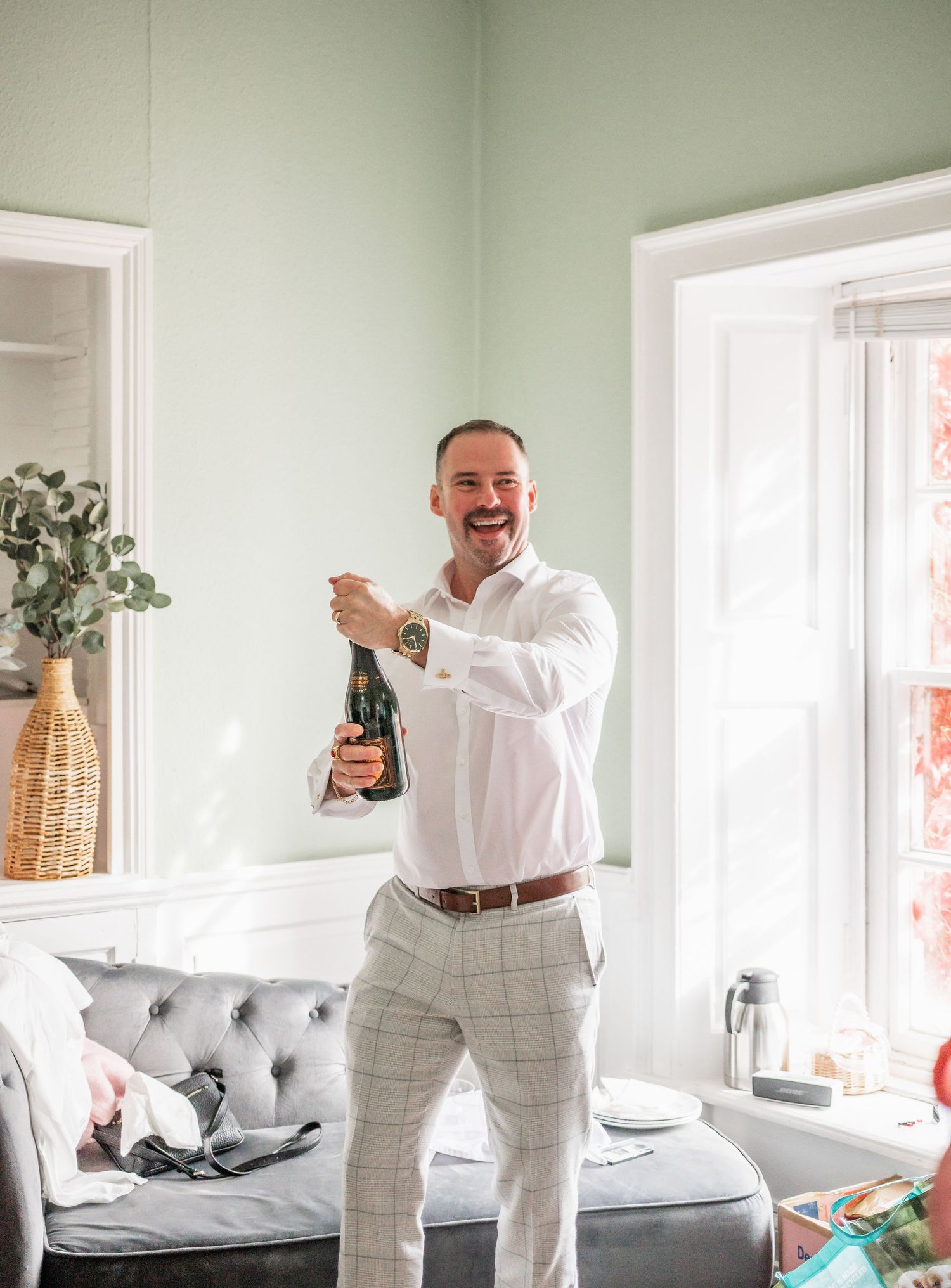 Person in white shirt and grey pants celebrating with champagne in a bright interior room.