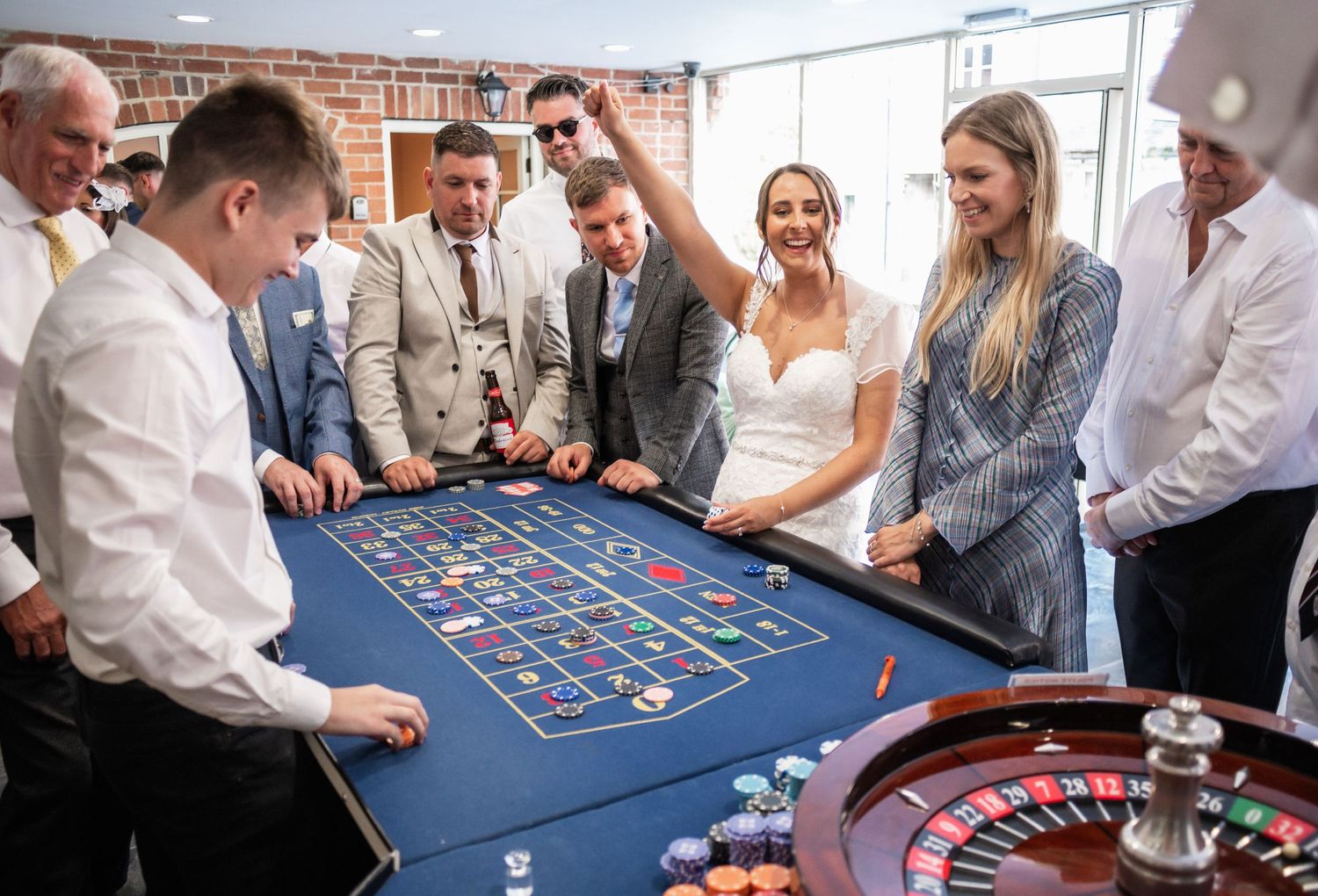 Wedding party gathered around blue roulette table for casino-themed entertainment.