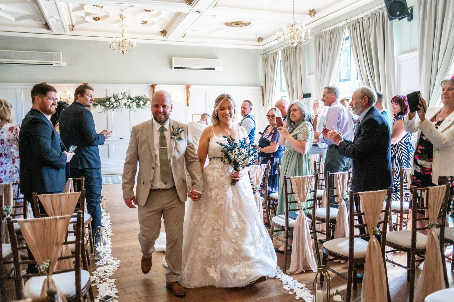 Wedding ceremony in an elegant room with chandelier as couple walks down aisle among seated guests.