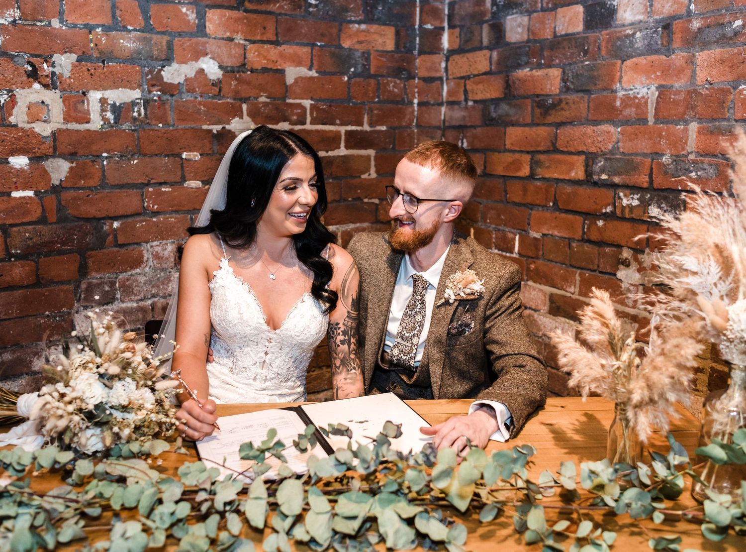 Newlyweds sit at decorated table with greenery and dried flowers against brick wall.