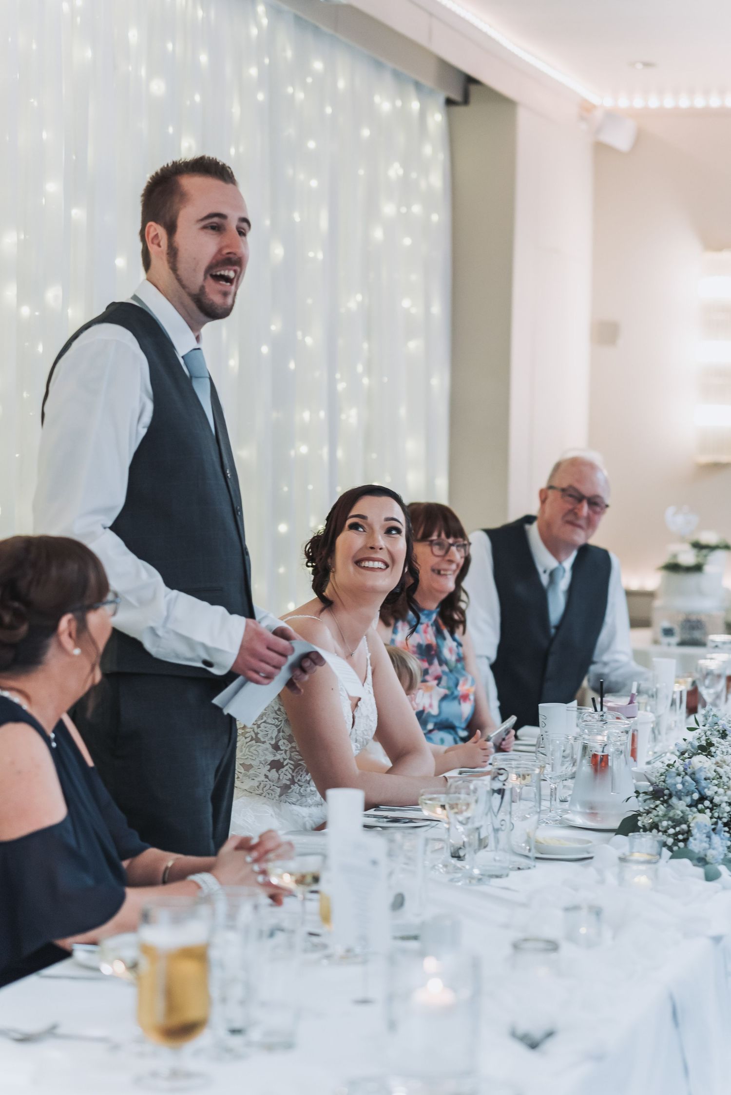 A wedding reception speech moment captured at a decorated table with twinkle light backdrop.