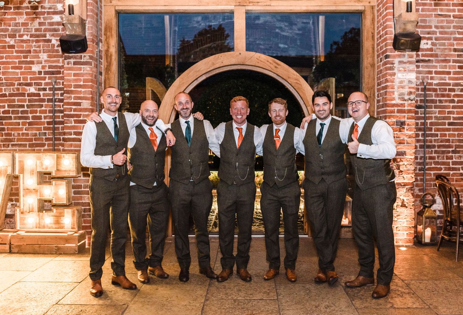 Groomsmen in matching vests pose together outside brick venue entrance.