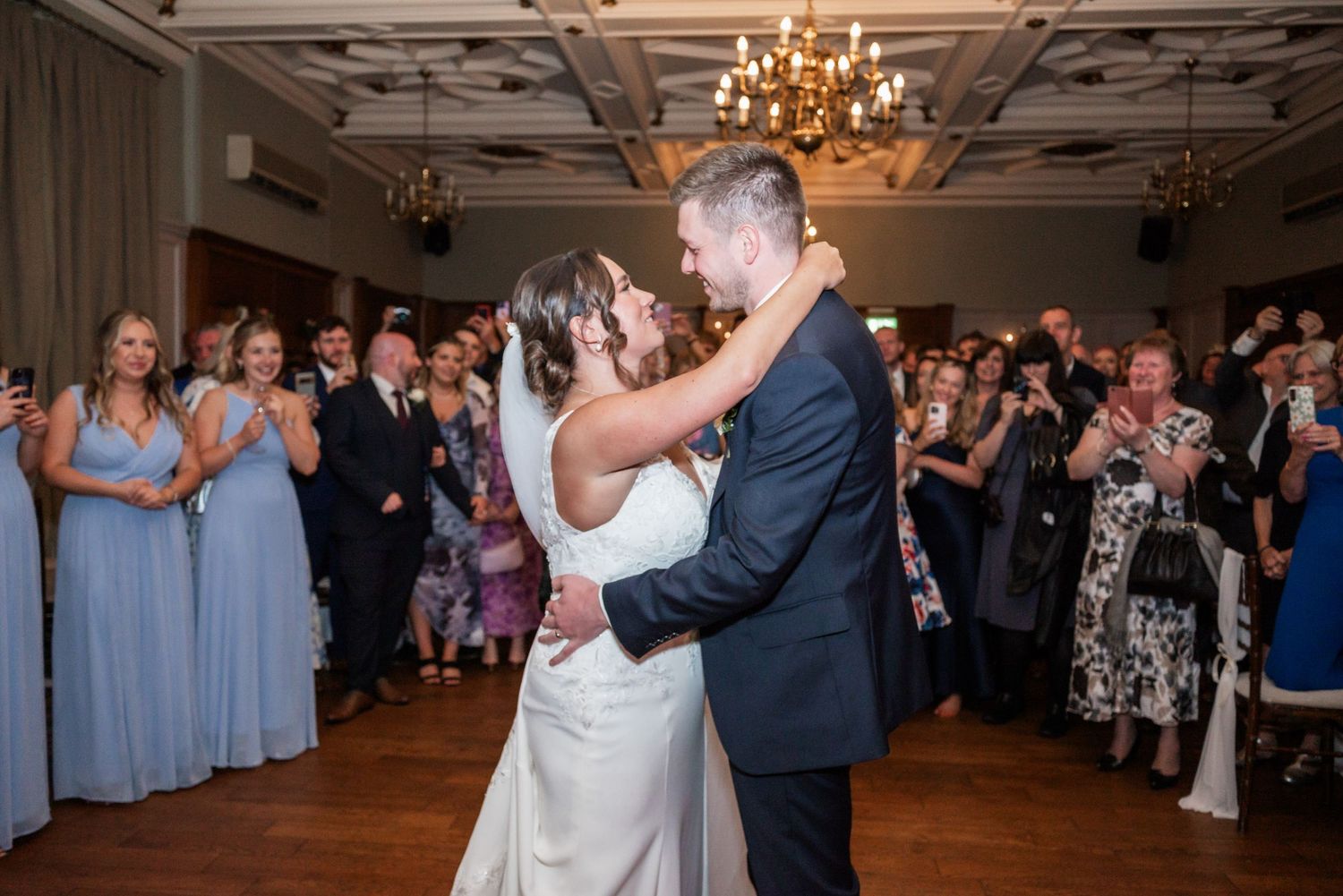 Couple shares their first dance surrounded by guests in an elegant ballroom with chandelier.
